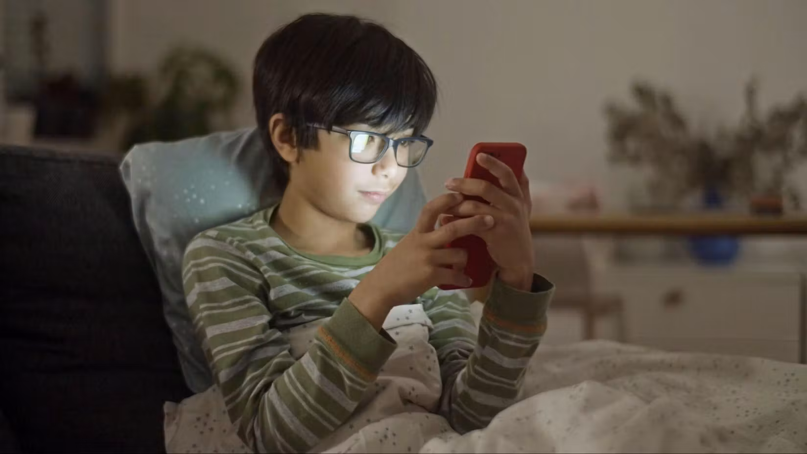 Photo of a child lying on a couch under a blanket, focused on a smartphone with a red case, in a cozy, softly lit room.