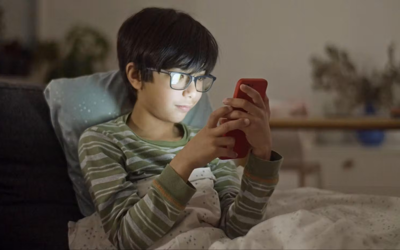 Photo of a child lying on a couch under a blanket, focused on a smartphone with a red case, in a cozy, softly lit room.