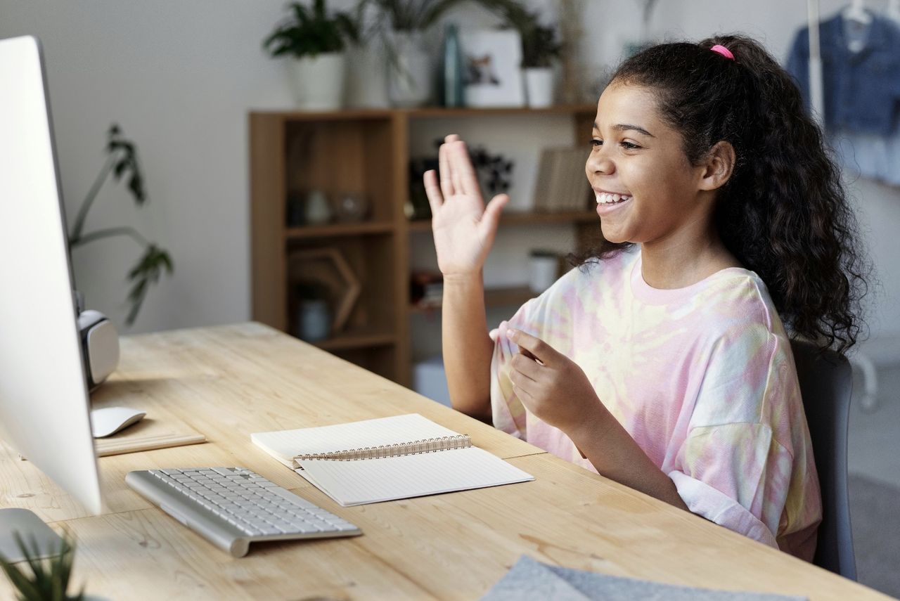 Photo of a smiling girl waving at a computer screen during an online coding lesson