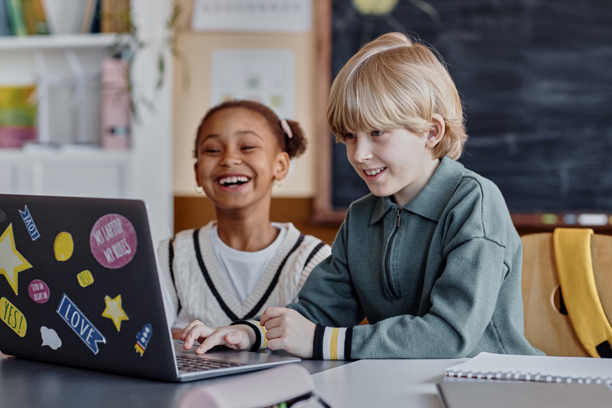 Photo of two children sitting at a desk, smiling and using a laptop decorated with colorful stickers in a classroom setting.