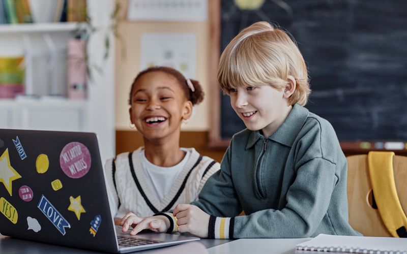 Photo of two children sitting at a desk, smiling and using a laptop decorated with colorful stickers in a classroom setting.