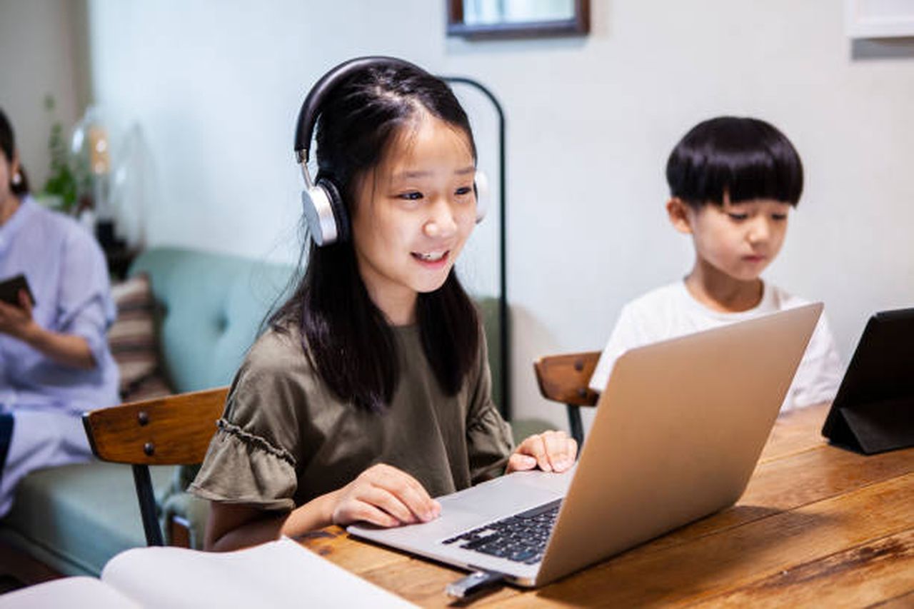 Photo of a girl wearing headphones using a laptop at home, sitting next to her brother