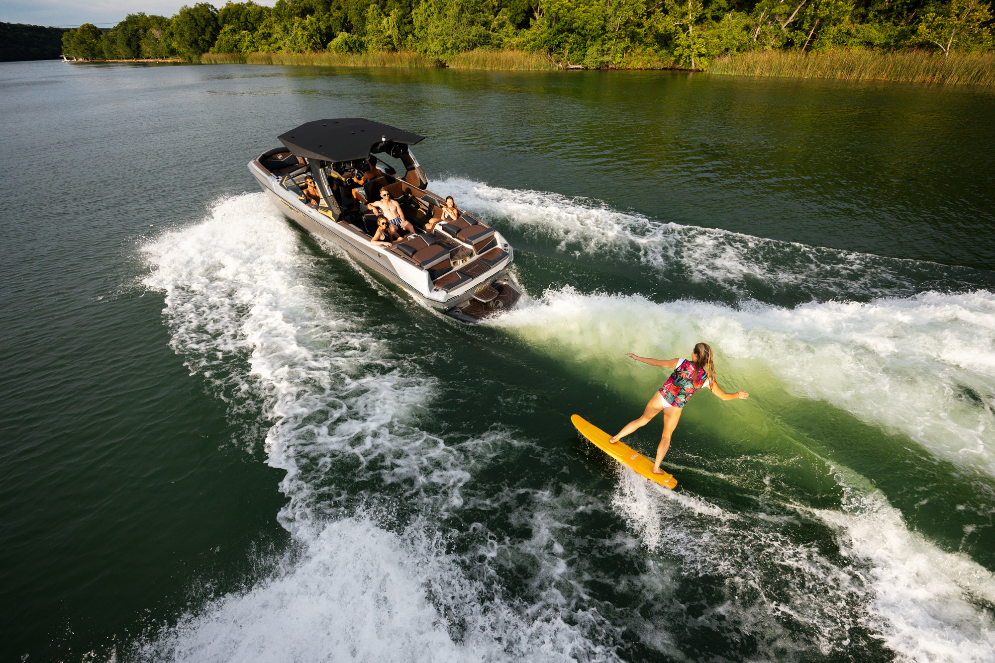 A person surfing behind a 2026 Tige Ultre 25ZX on a calm lake, surrounded by lush green hills.