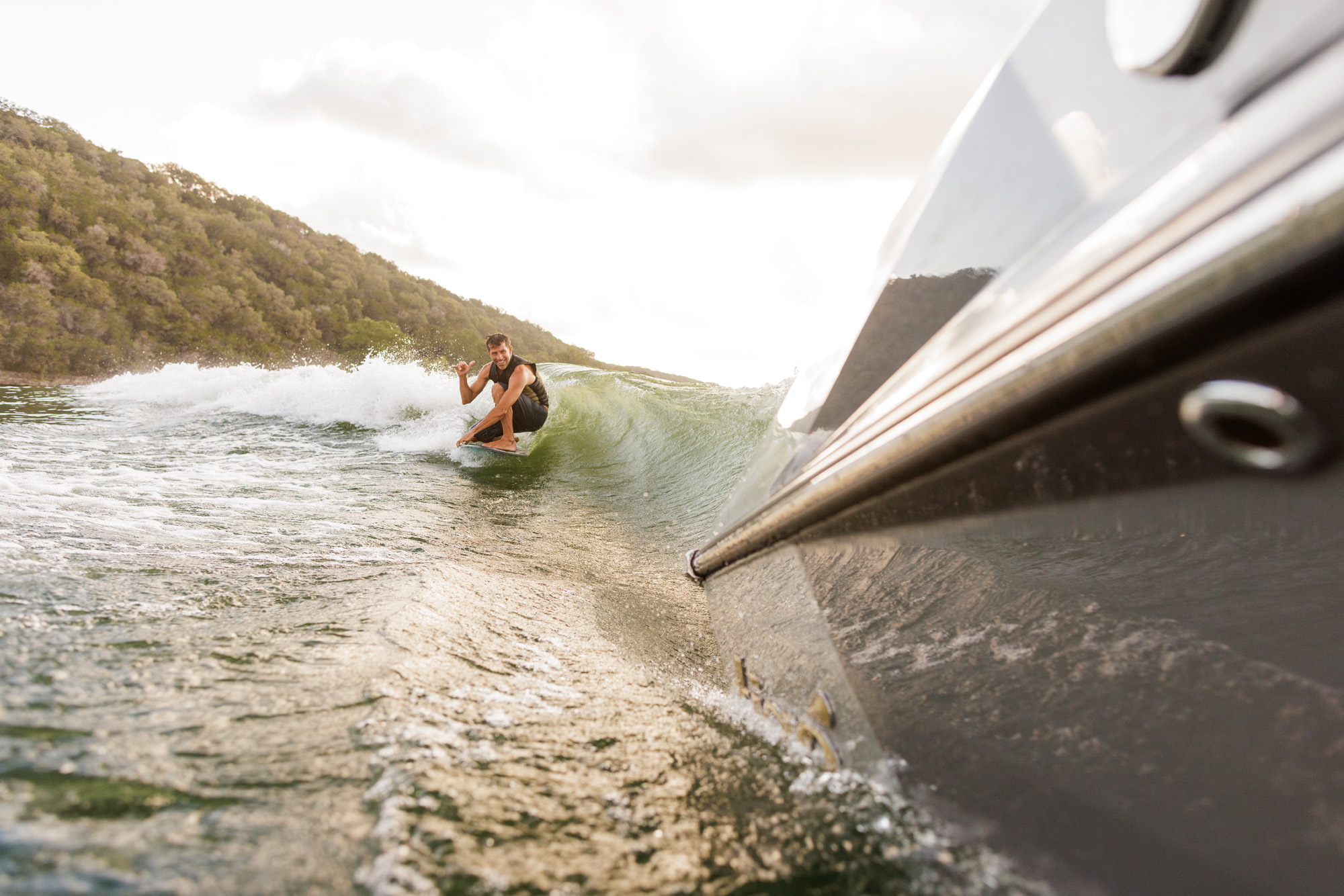 A person surfing behind a 2026 Tige Ultre 25ZX boat, surrounded by green hills under a cloudy sky.