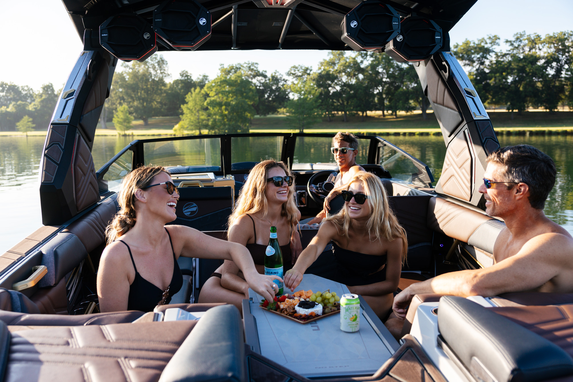 Five people in swimsuits enjoy snacks and drinks on the RetracTable on the all new 2026 Tige Ultre 25ZX boat, surrounded by water and trees, under a clear sky.