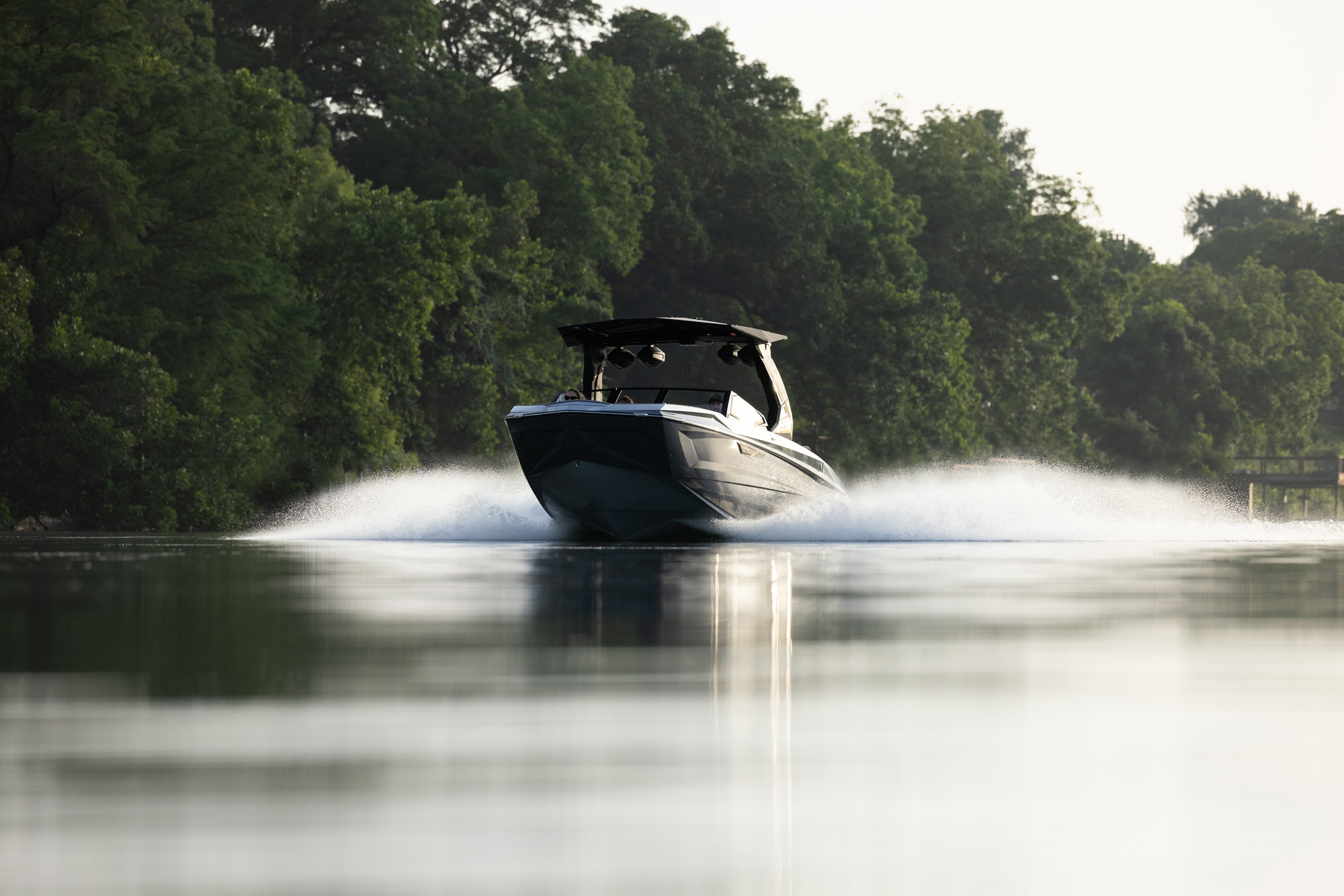 A 2026 Tige Ultre 25ZX swiftly moves across a calm lake, creating a wake. Lush green trees line the background under a clear sky.