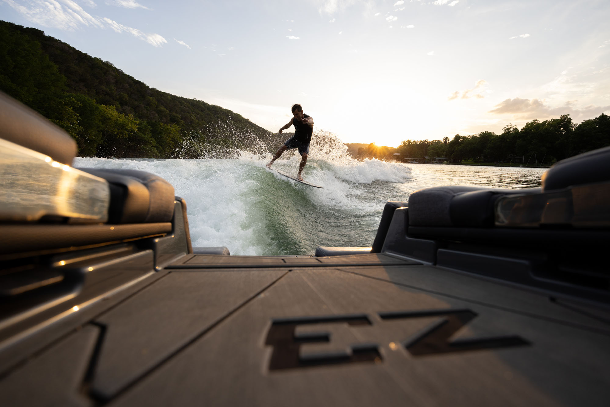 Person surfing on a lake at sunset, viewed from a 2026 Tige Z3 boat. Forested hills and a bright sky are in the background.