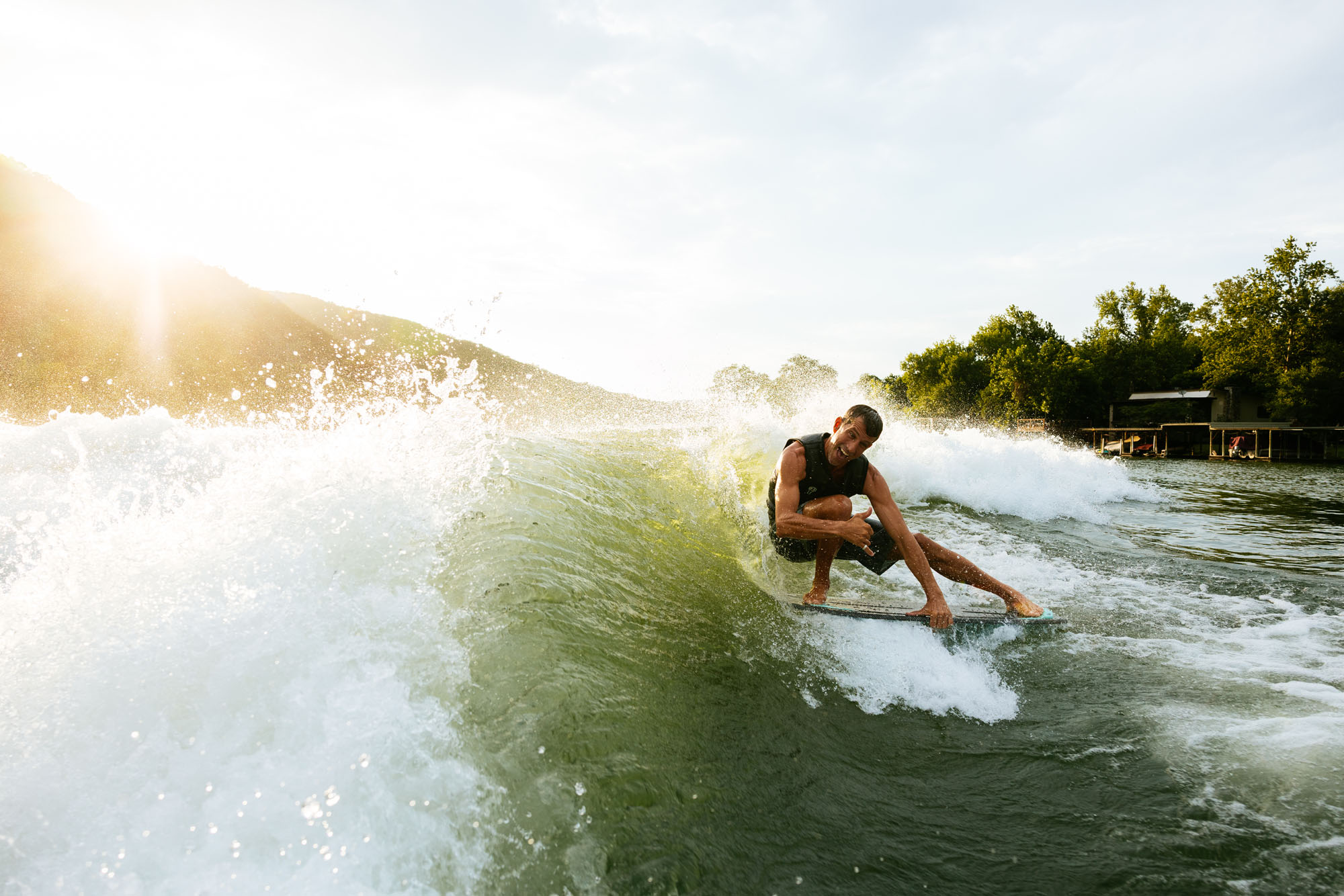 A person surfing on a wave behind a 2026 Tige Z3 during sunset, surrounded by splashing water, with trees and a small building in the background.