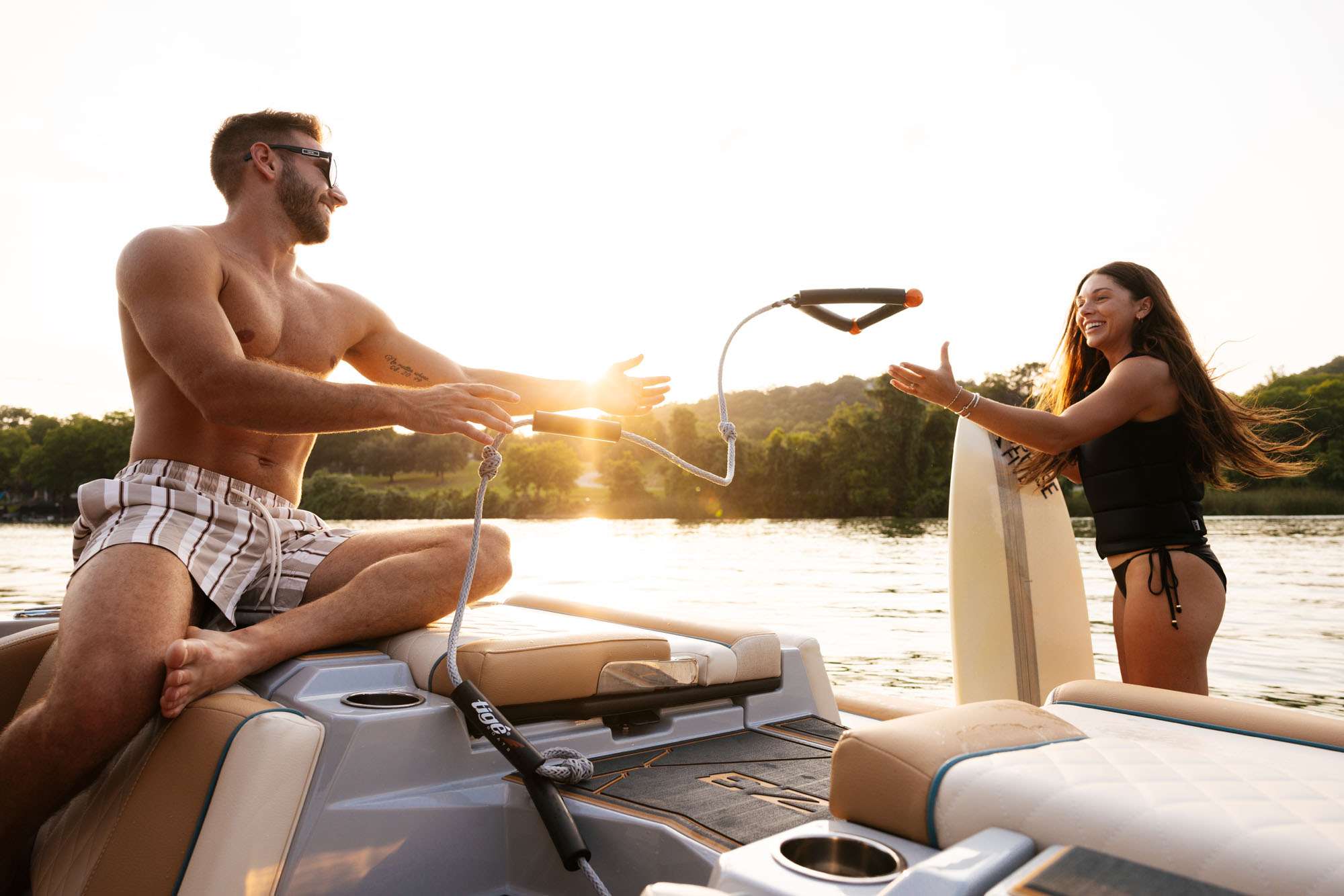 A man sitting on the transom of a 2026 Tige Z3 boat hands a surf rope to a woman in a life vest, with a sunset in the background over a lake.