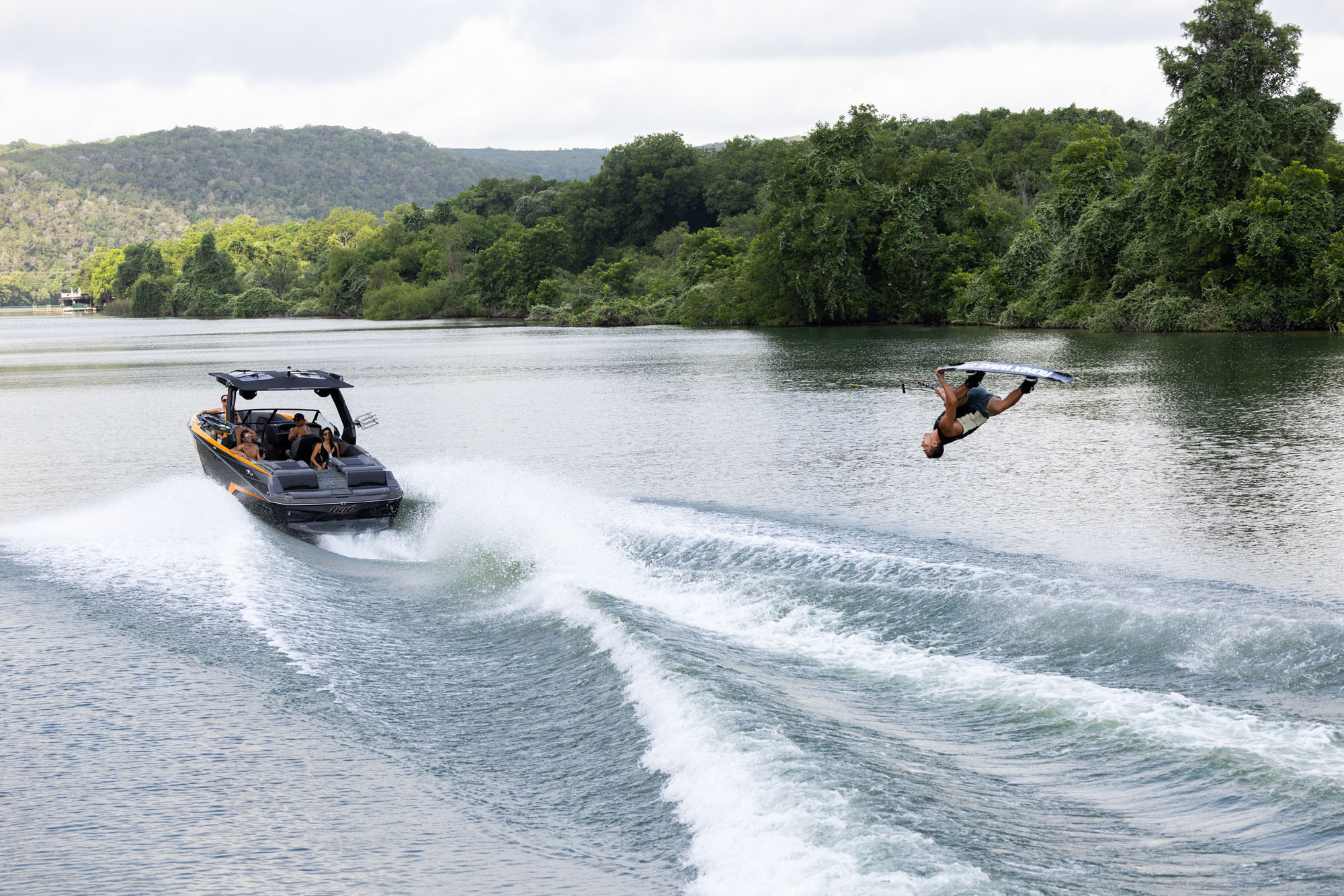A wakeboarder performs an aerial trick behind a speeding 2026 Tige Z3 boat on Lake Austin, surrounded by lush green hills and trees.