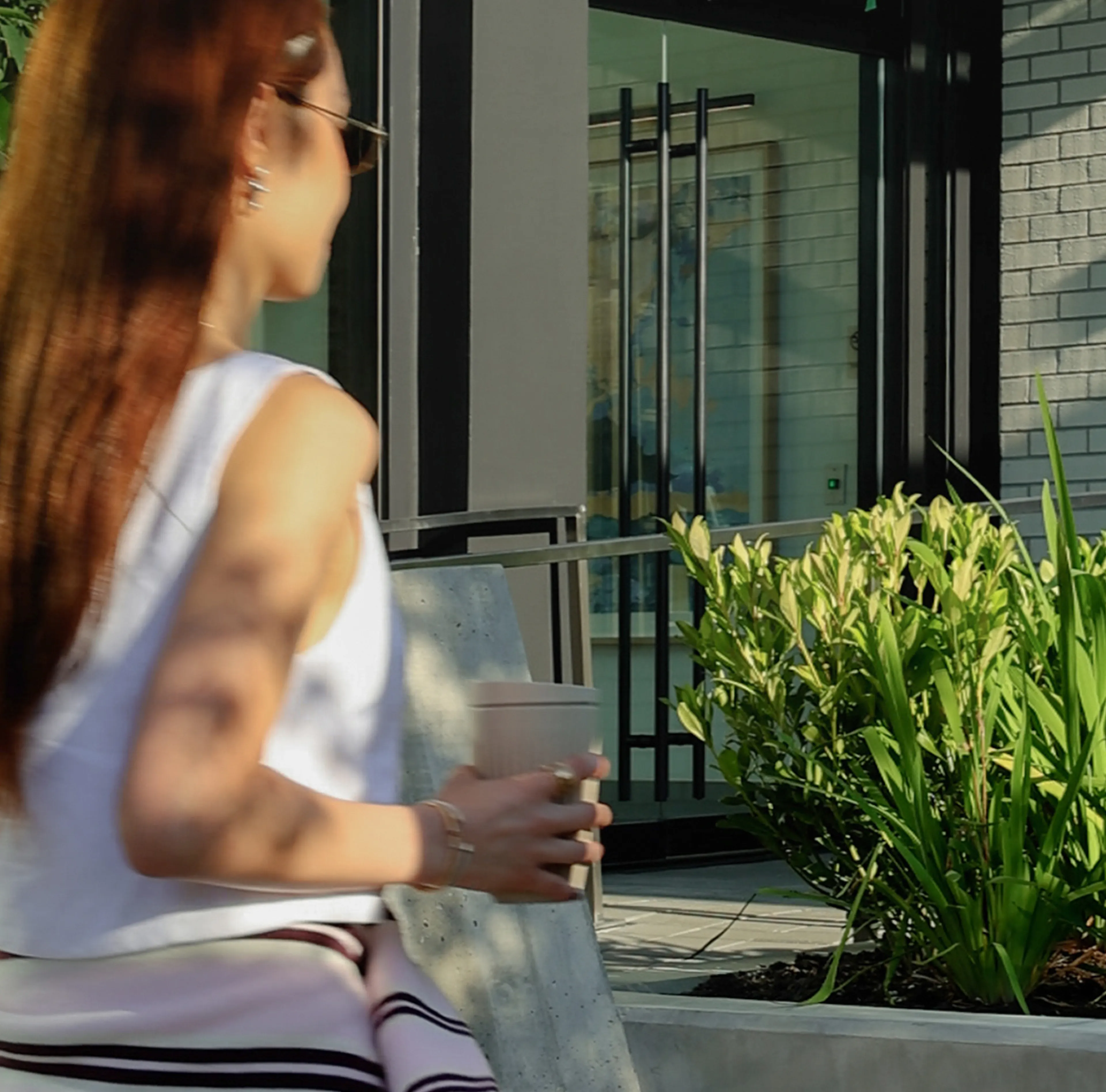 A woman with long hair walks outside a modern building holding a drink, with greenery and sunlight casting shadows around her.