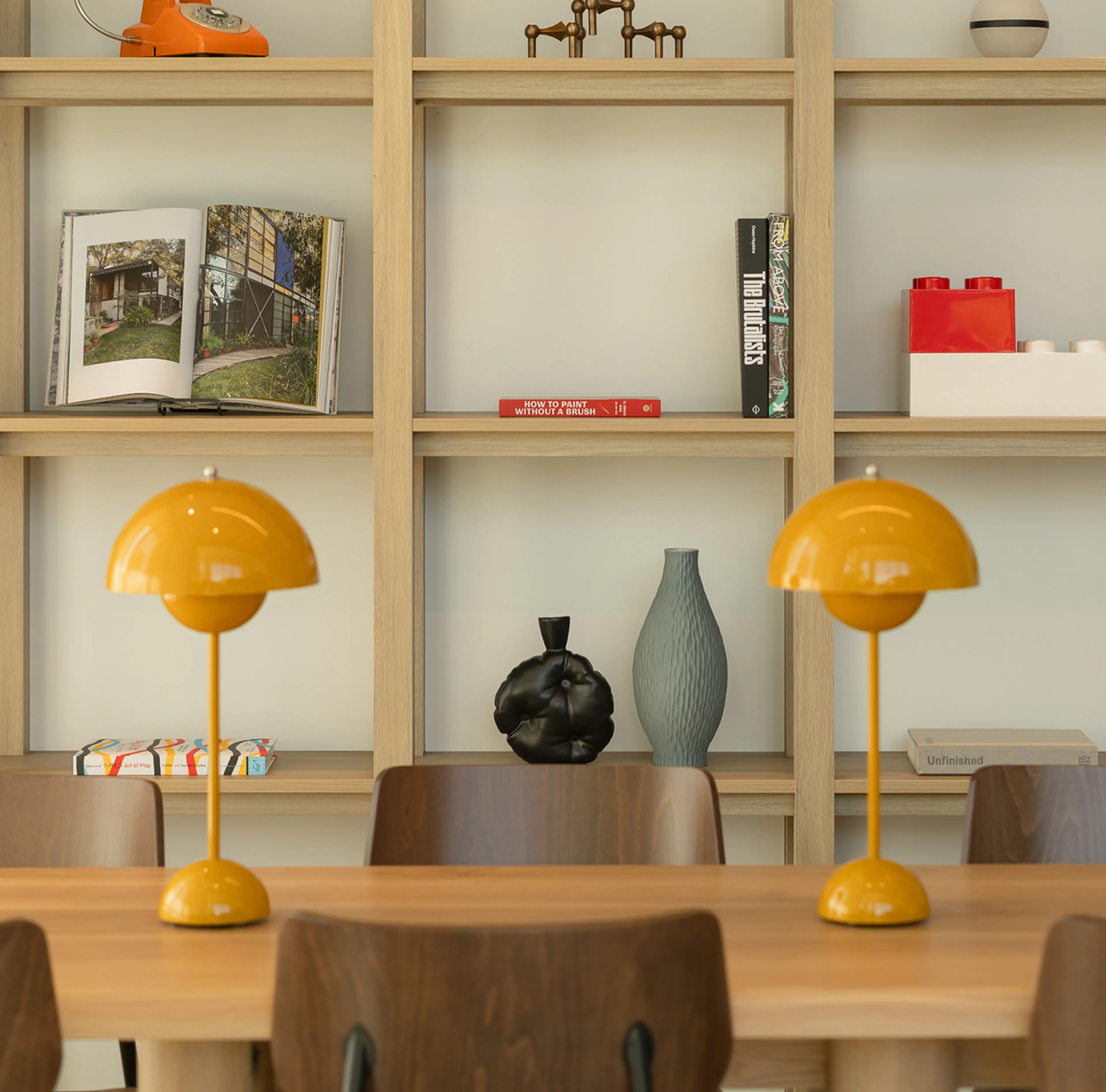 A modern dining area with a wooden table, yellow lamps, brown chairs, and a light wooden bookshelf displaying books, vases, and decor items.