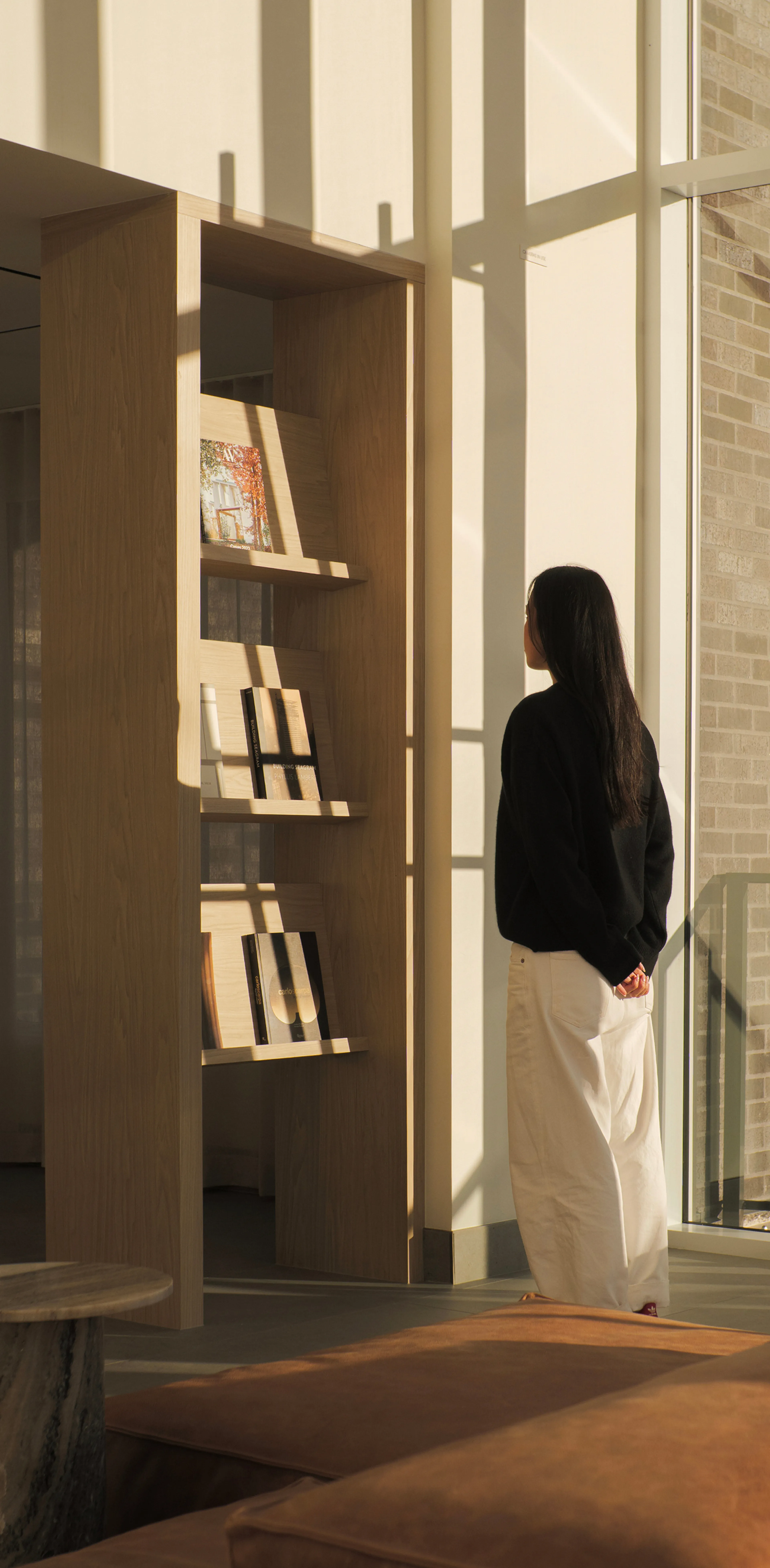 A person in a black top and white pants stands in front of a wooden bookshelf, lit by soft sunlight, casting shadows on the wall.