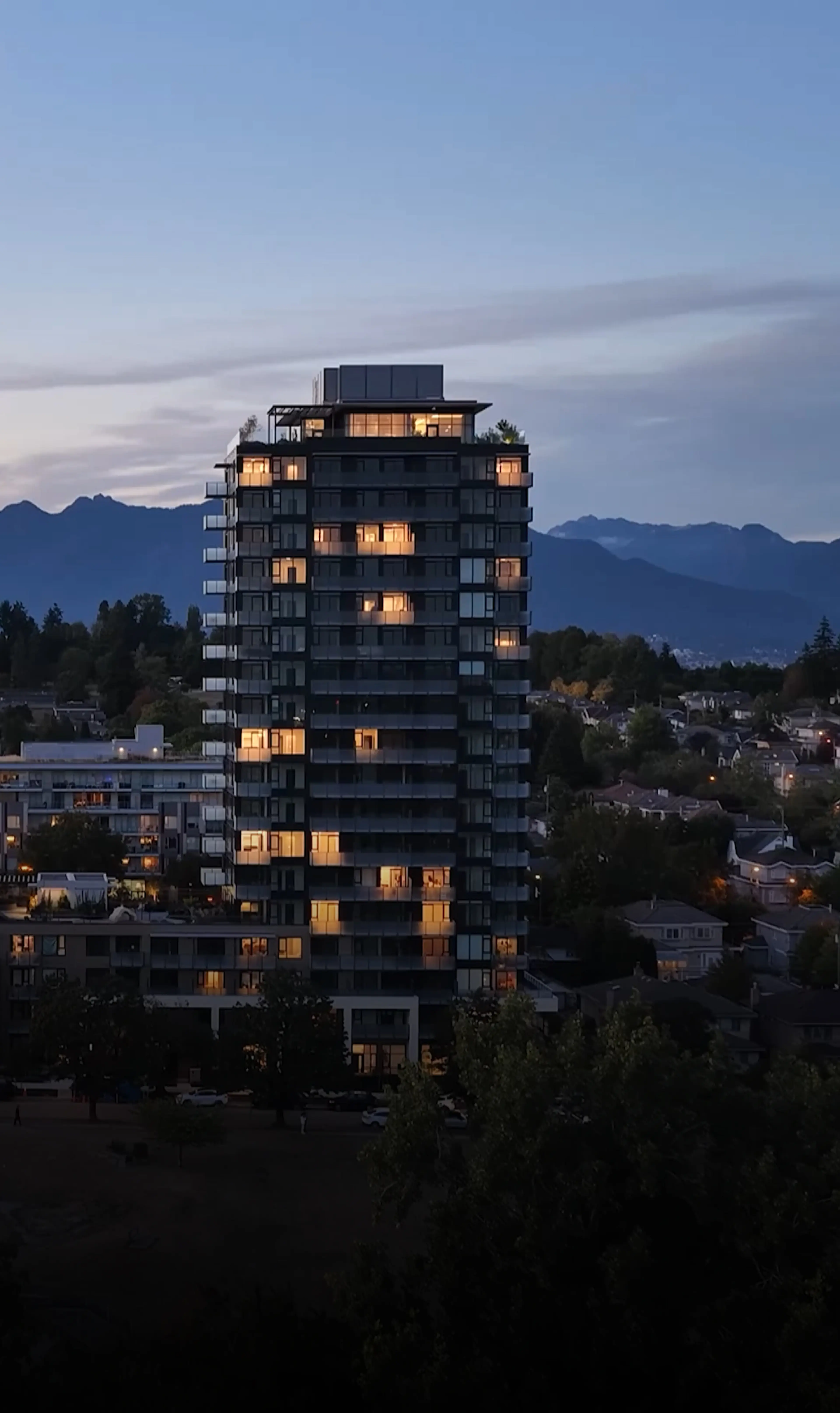 Tall building with illuminated windows at dusk, surrounded by trees and smaller buildings, set against a backdrop of distant mountains.