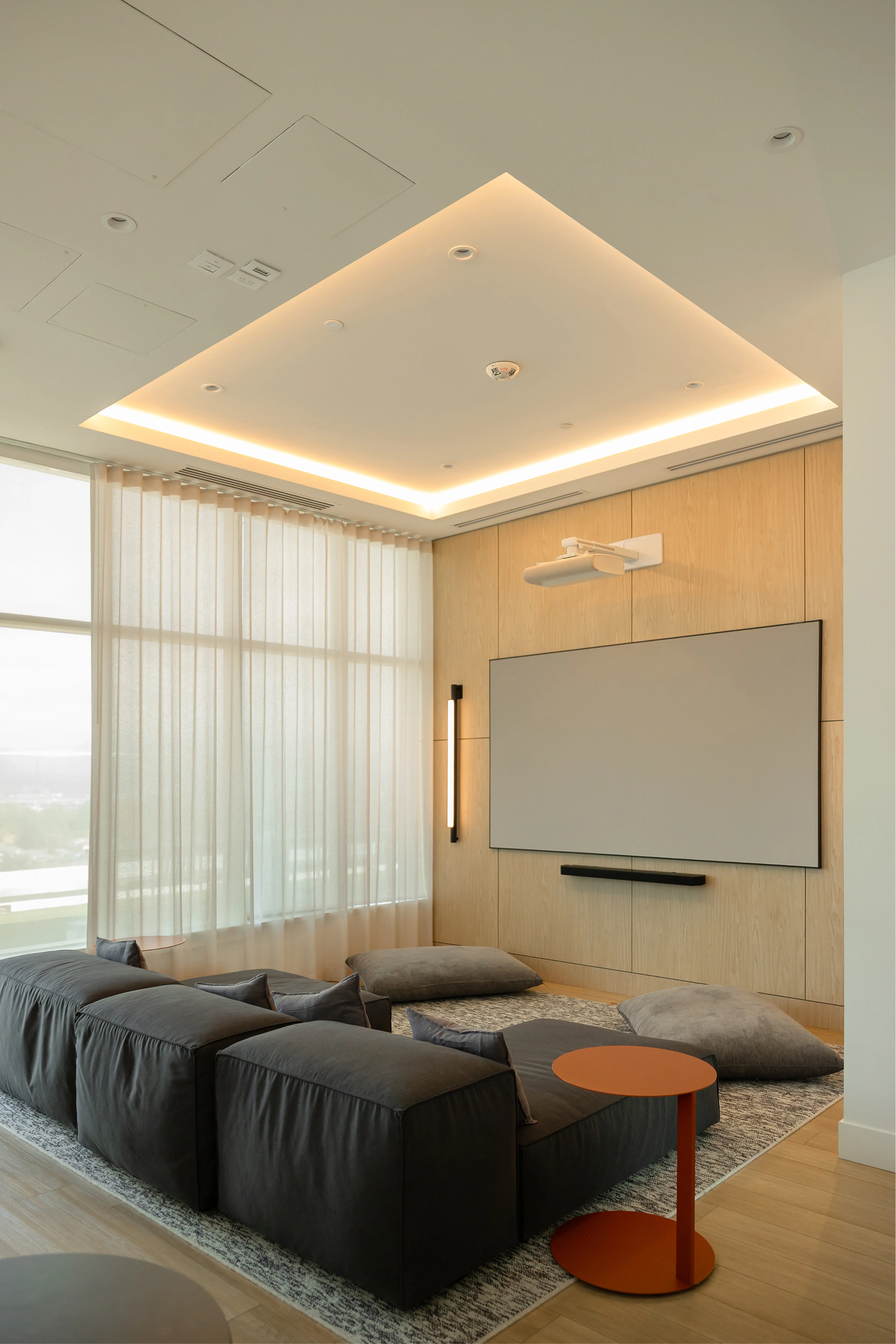 Modern living room with dark gray modular seating, large screen, wood paneling, and soft lighting. Light curtains cover tall windows.