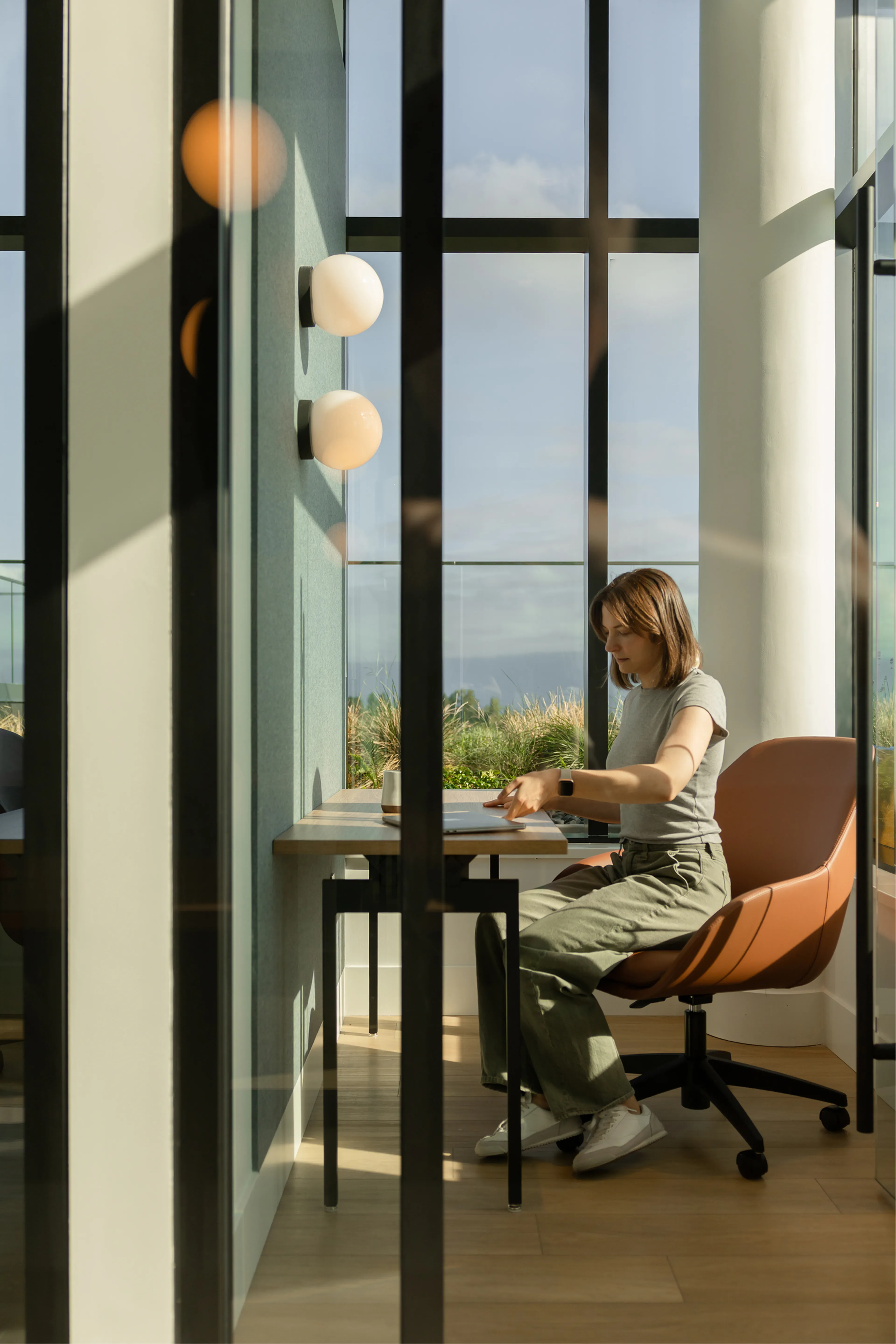 A woman in a sunlit office room sits at a desk, working on a laptop. She is wearing casual clothes, and large windows reveal a view of greenery outside.