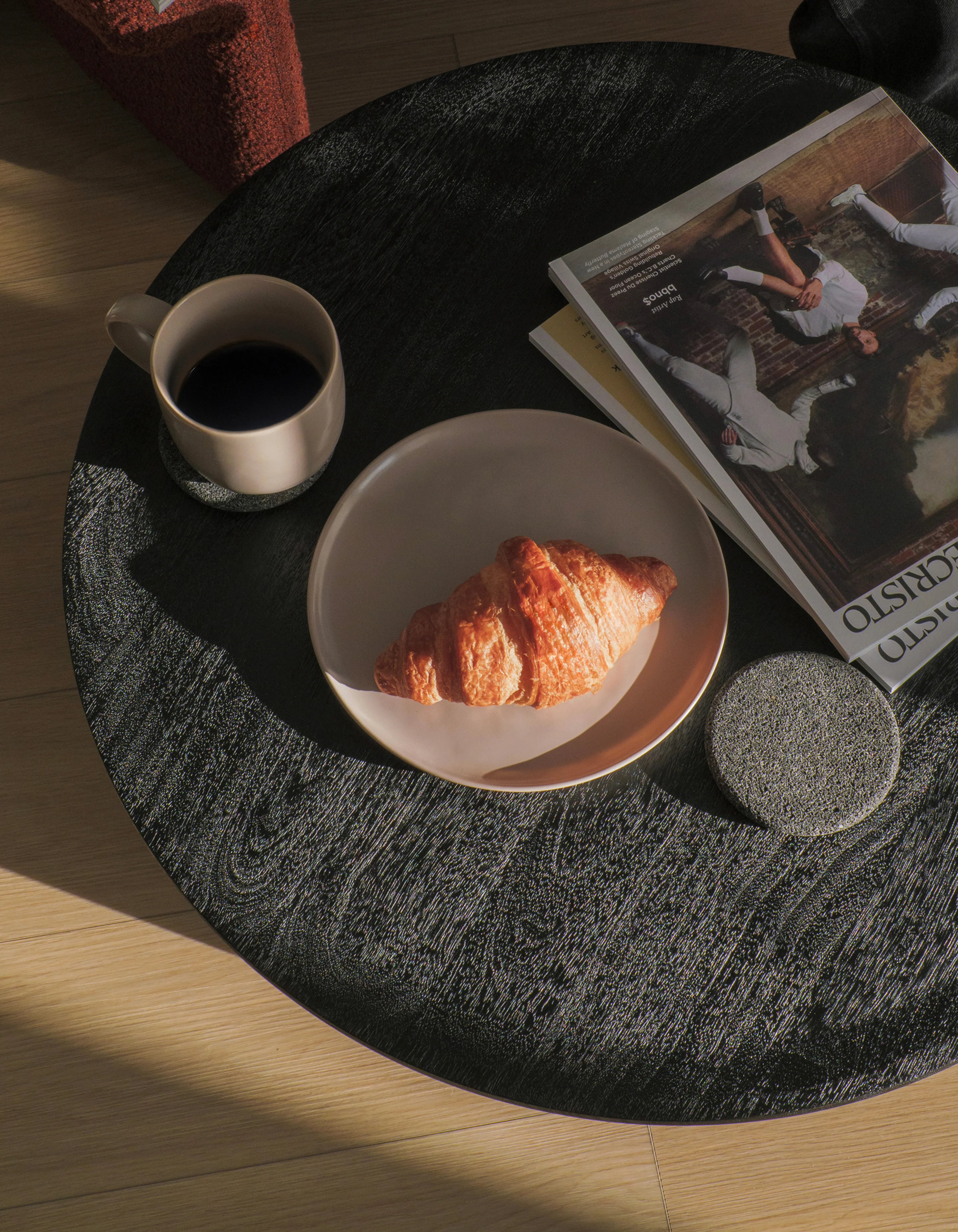 A croissant on a plate, a cup of coffee, and magazines on a round black table, lit by warm sunlight.