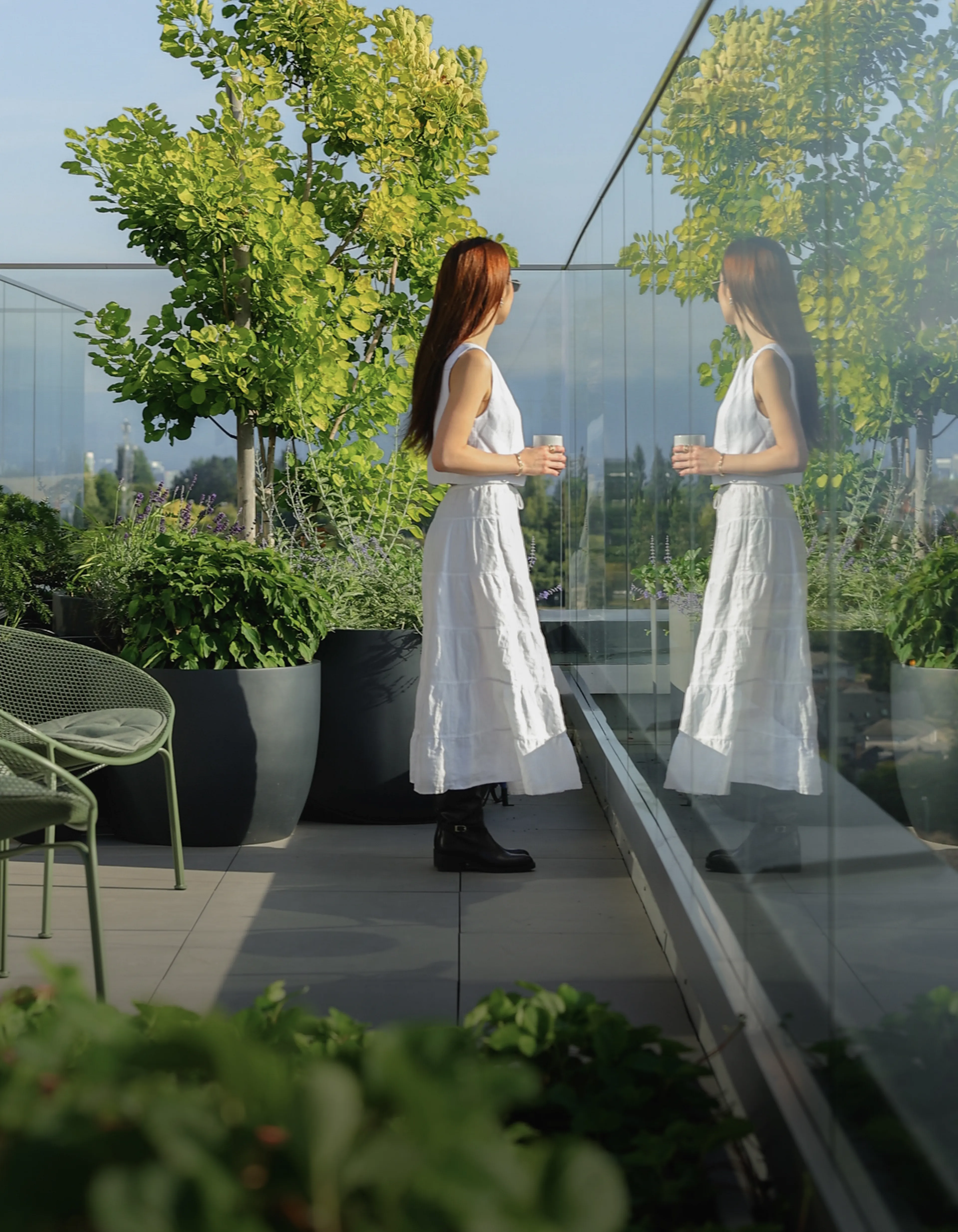 A woman in a white dress holding a cup, standing on a terrace with green plants and trees, her reflection visible in a glass wall.