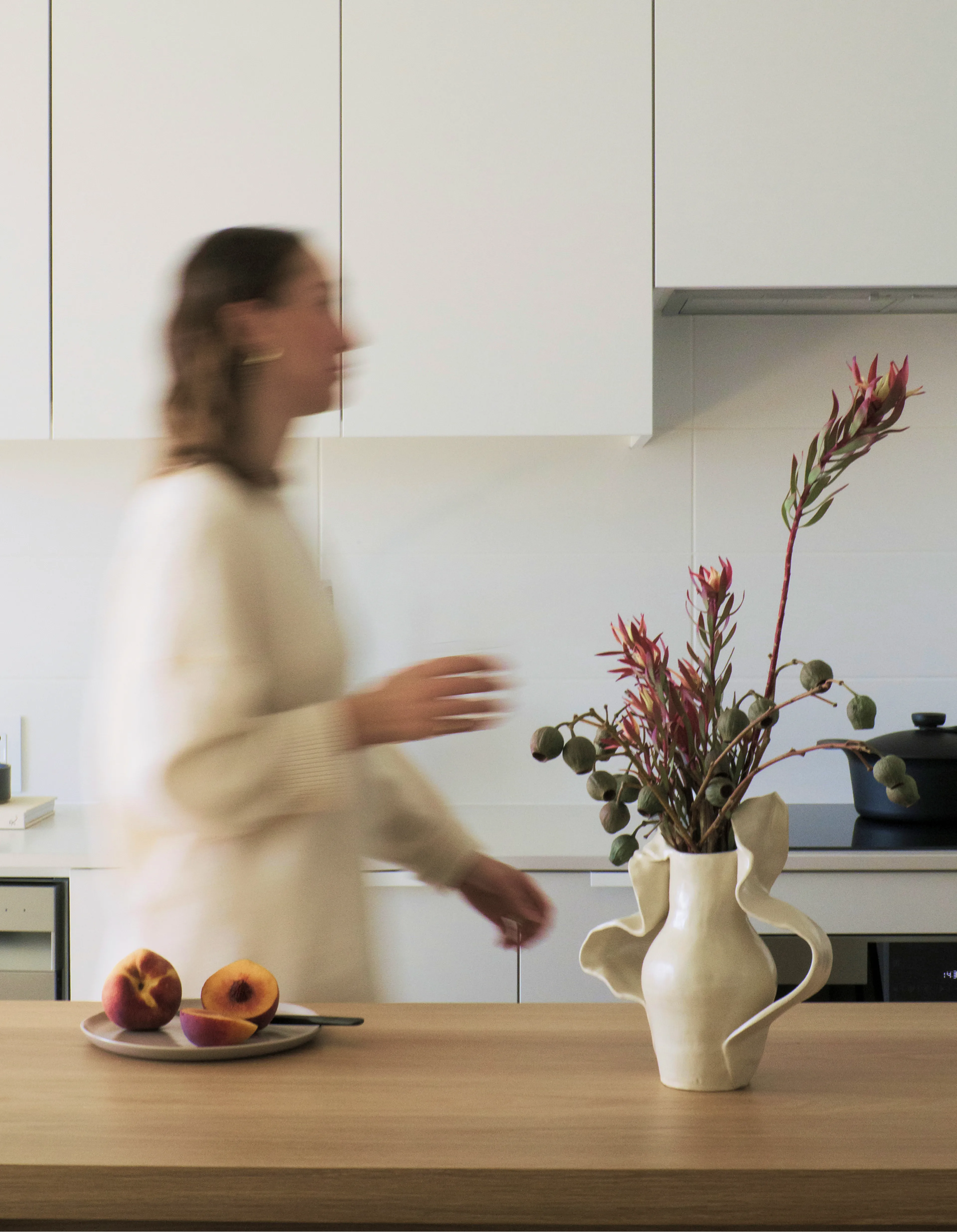 A person in motion blurs past a kitchen counter with a ceramic vase of flowers and a plate of peaches.