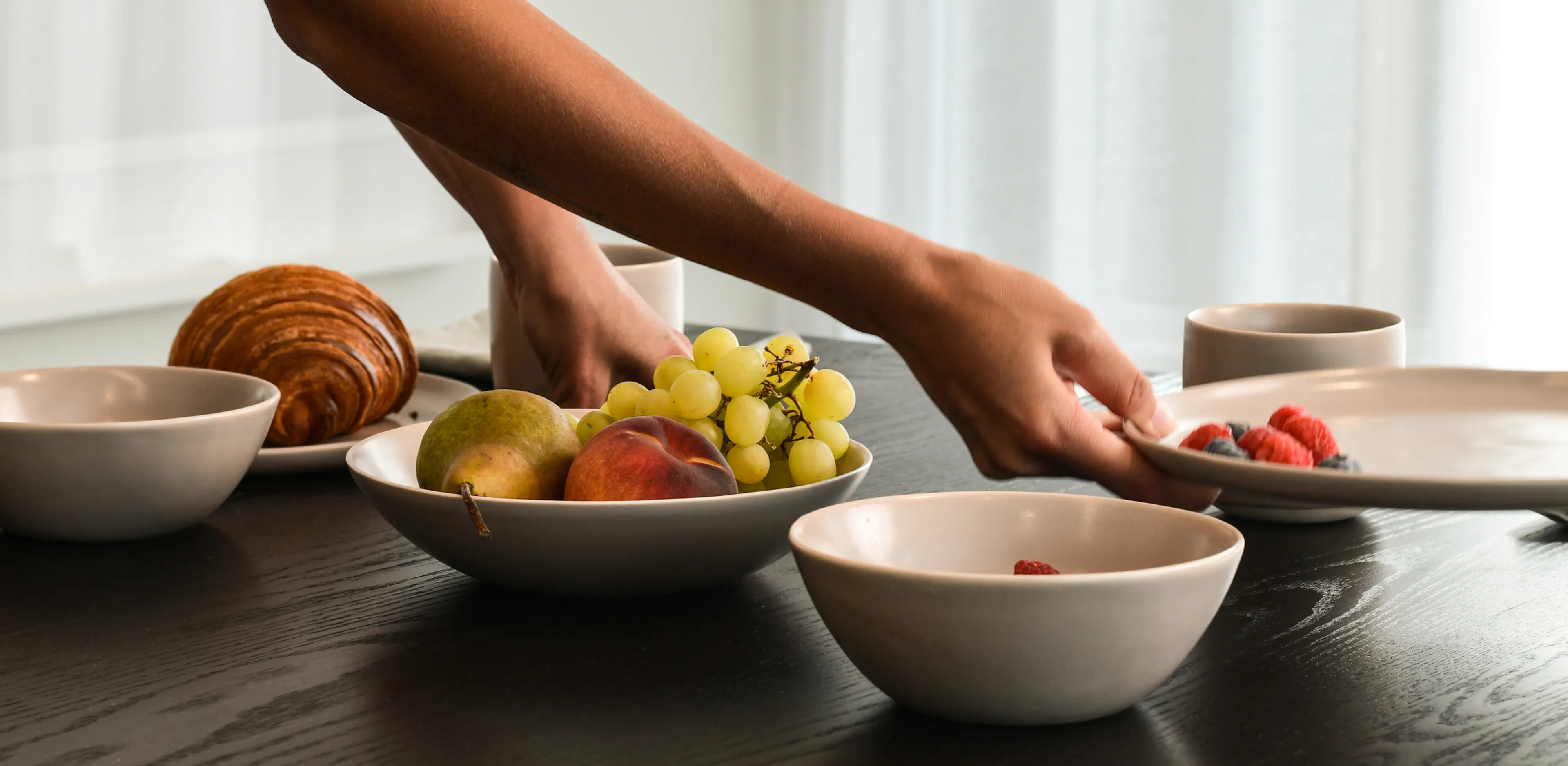 A person setting a table with dishes containing green grapes, a pear, a peach, a croissant, and a plate of raspberries and blueberries.