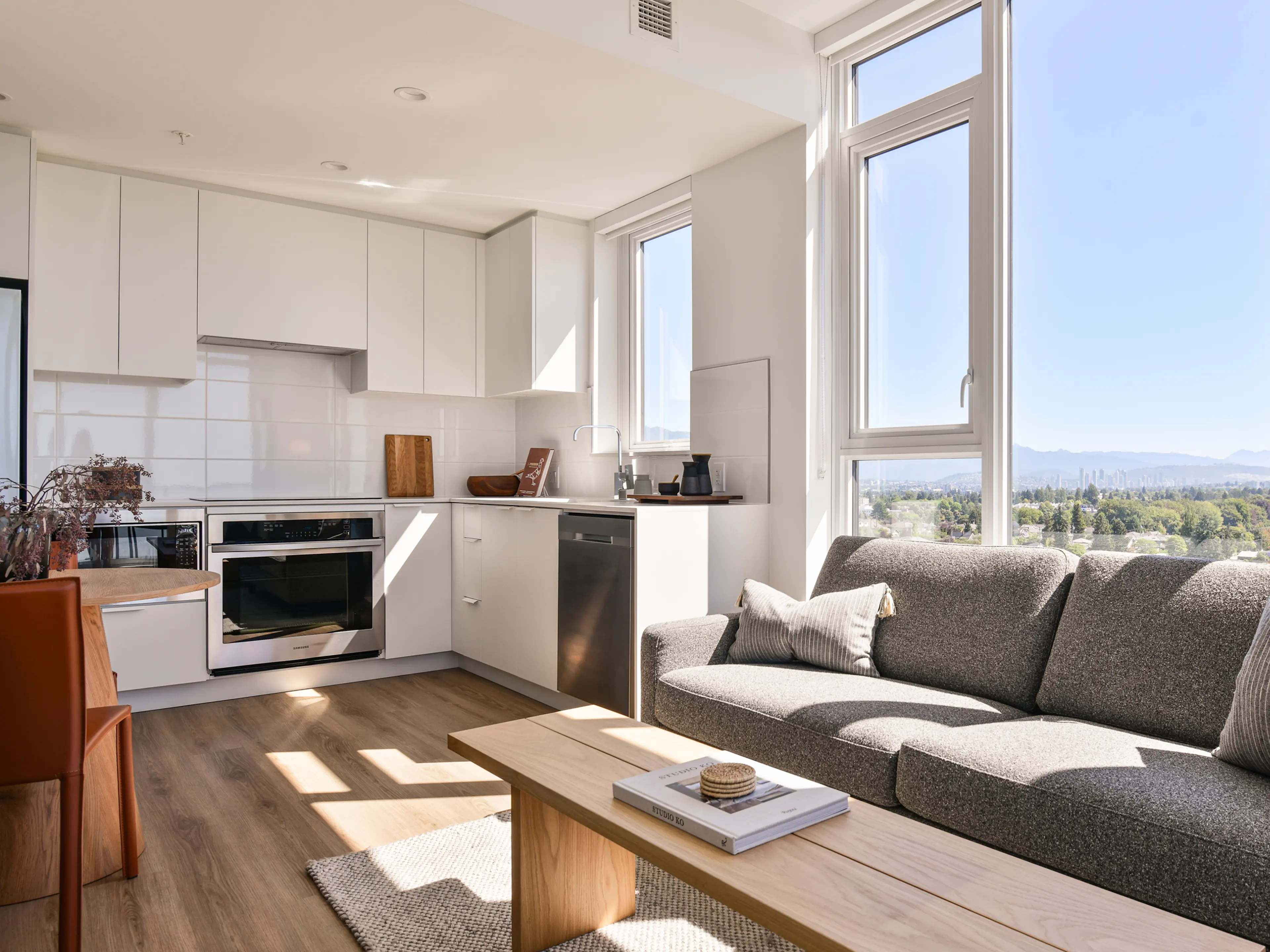 Modern, sunlit open-plan living room and kitchen with white cabinets, gray sofa, wooden floor, and large windows with a scenic view outside.