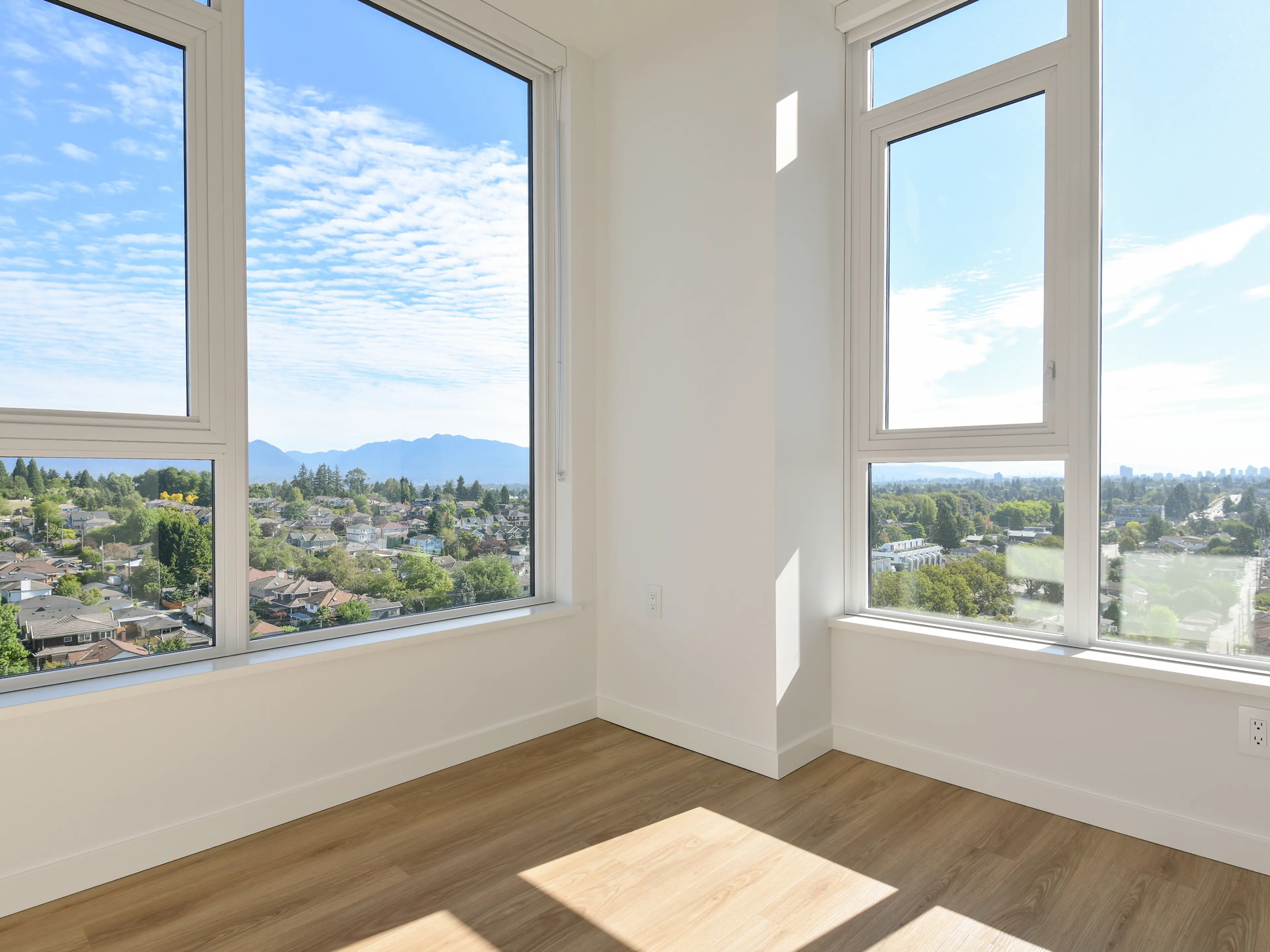 Bright, empty room with hardwood floors and large windows offering a scenic view of a suburban neighborhood and distant mountains.