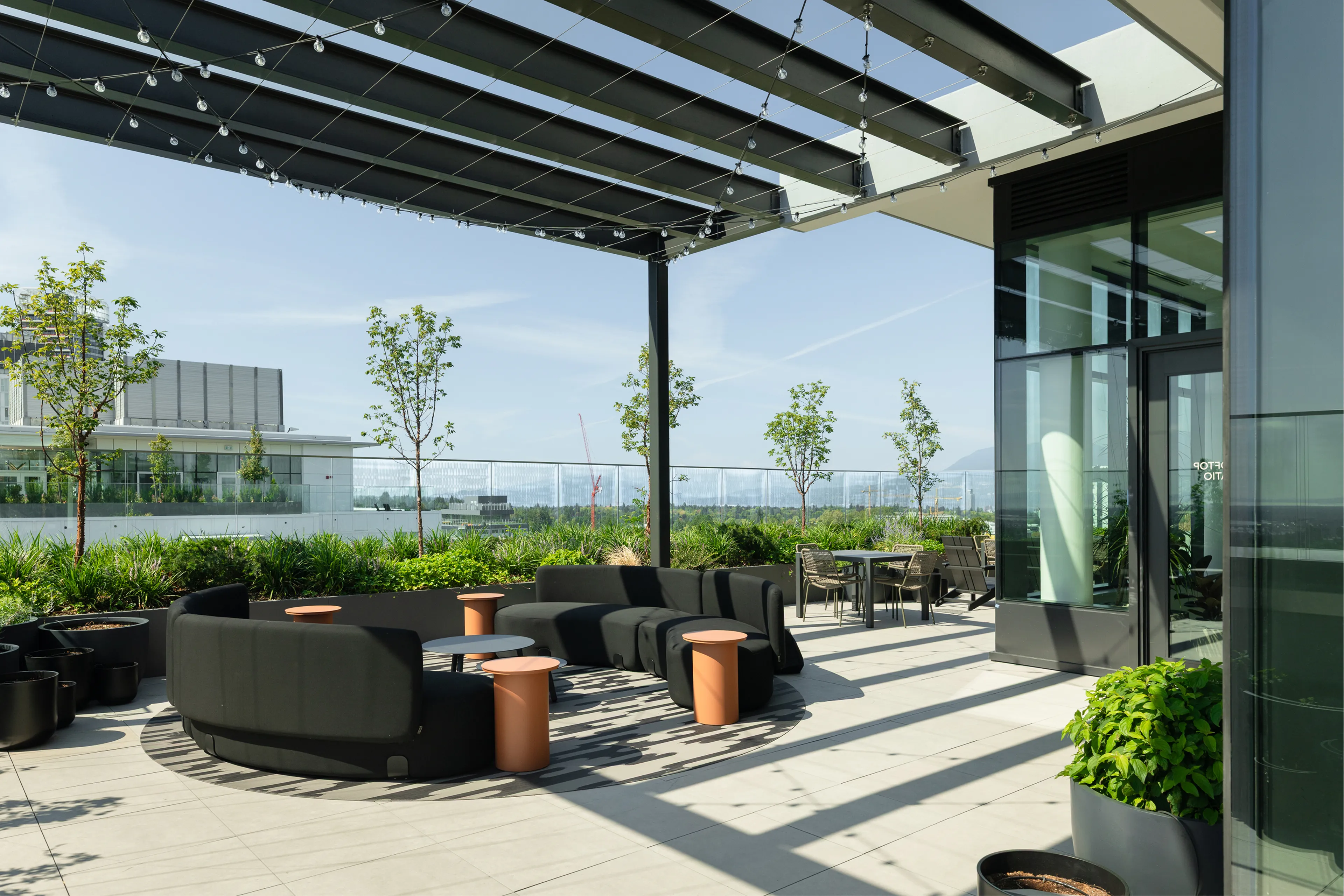 A modern rooftop terrace with black sofas, circular tables, and potted plants under a pergola, overlooking city buildings under a clear sky.