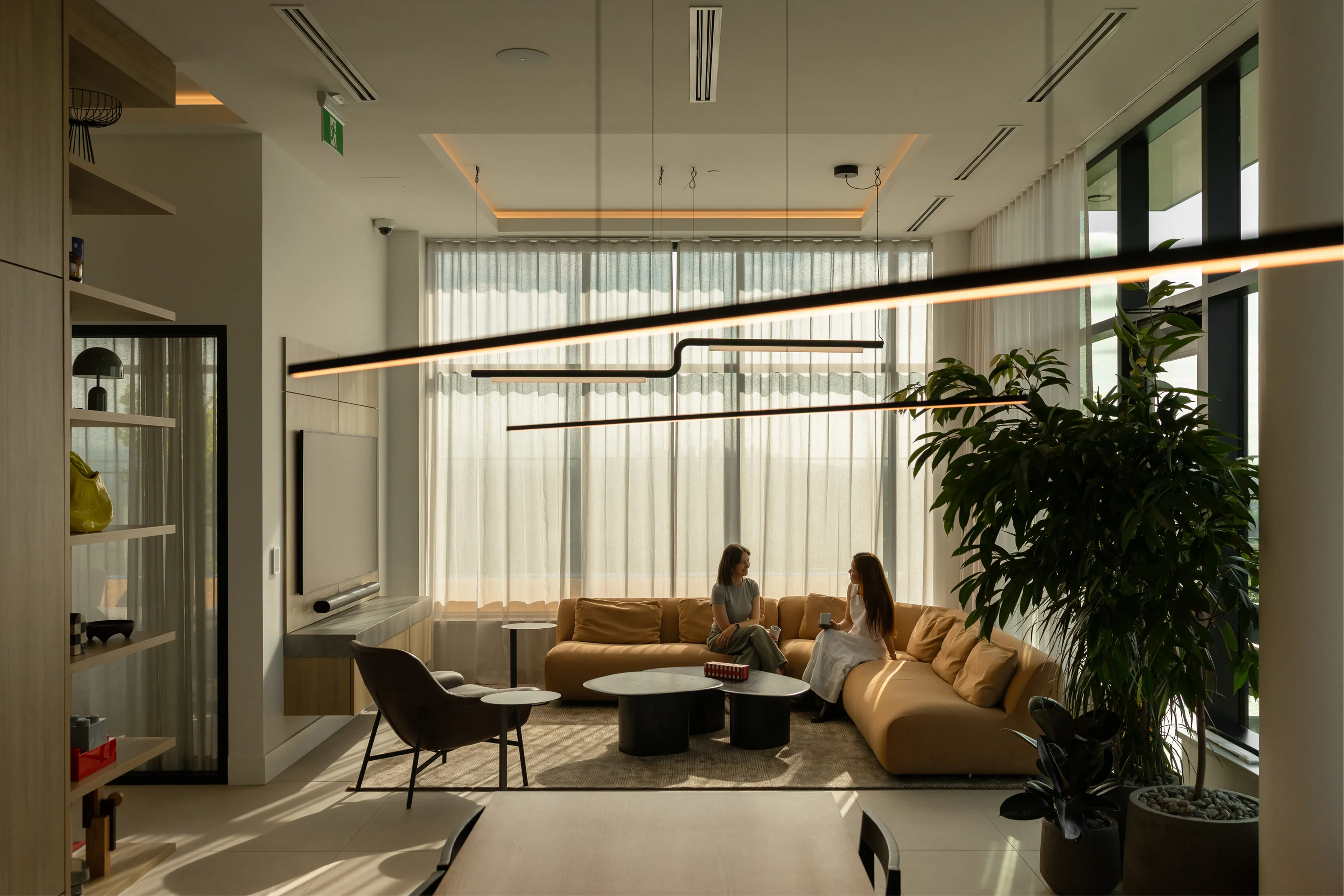 Two people sitting on a tan sectional sofa in a modern, well-lit living room with large windows and indoor plants.