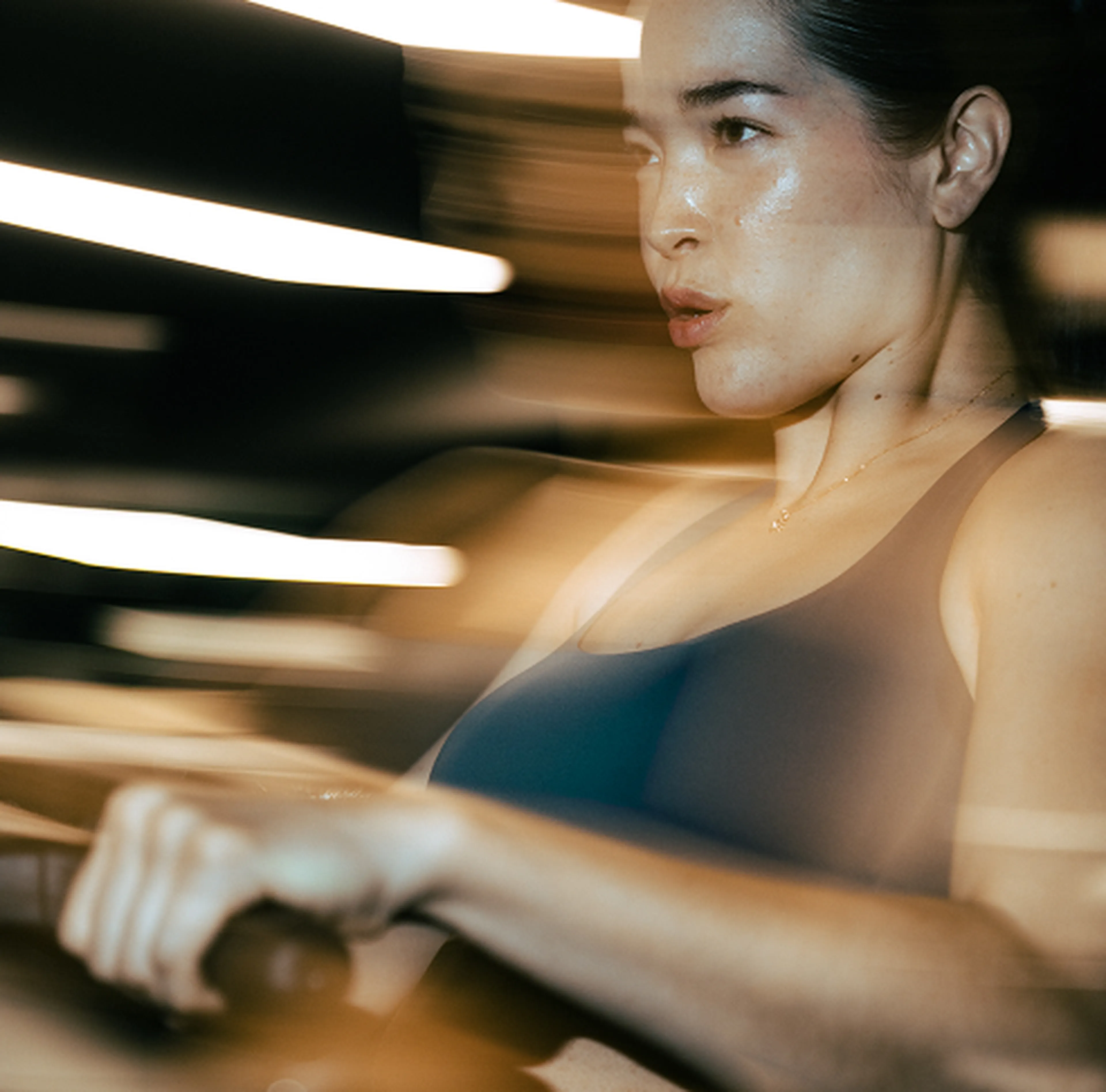 Woman intensely exercising on a rowing machine, with dynamic motion blur and focused expression, in a dimly lit gym environment.