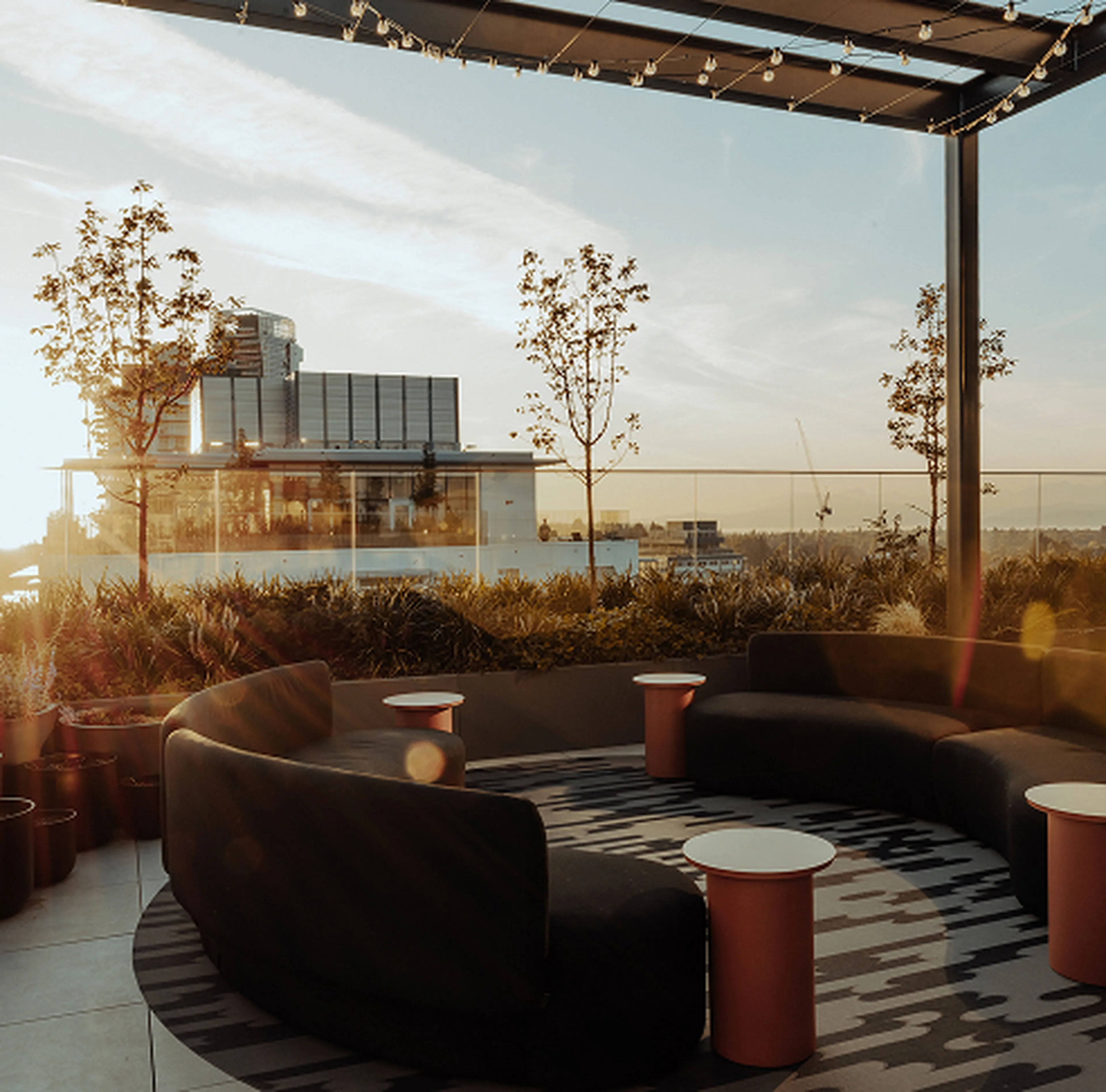Modern rooftop patio with curved seating, small tables, plants, and a view of a cityscape at sunset. String lights hang overhead.