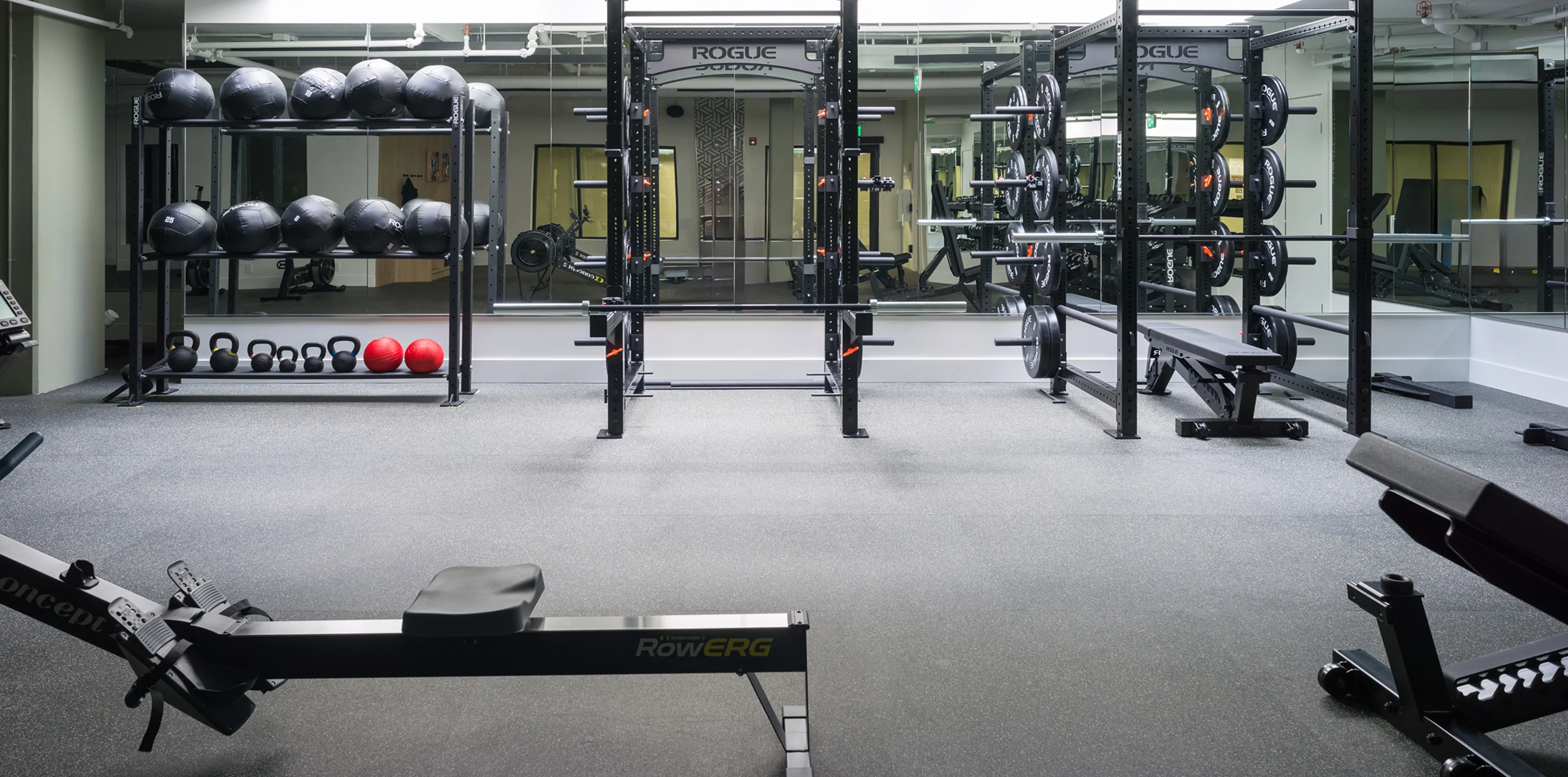 A modern gym with squat racks, weight plates, kettlebells, medicine balls, and a rowing machine. Large mirrors cover the back wall.