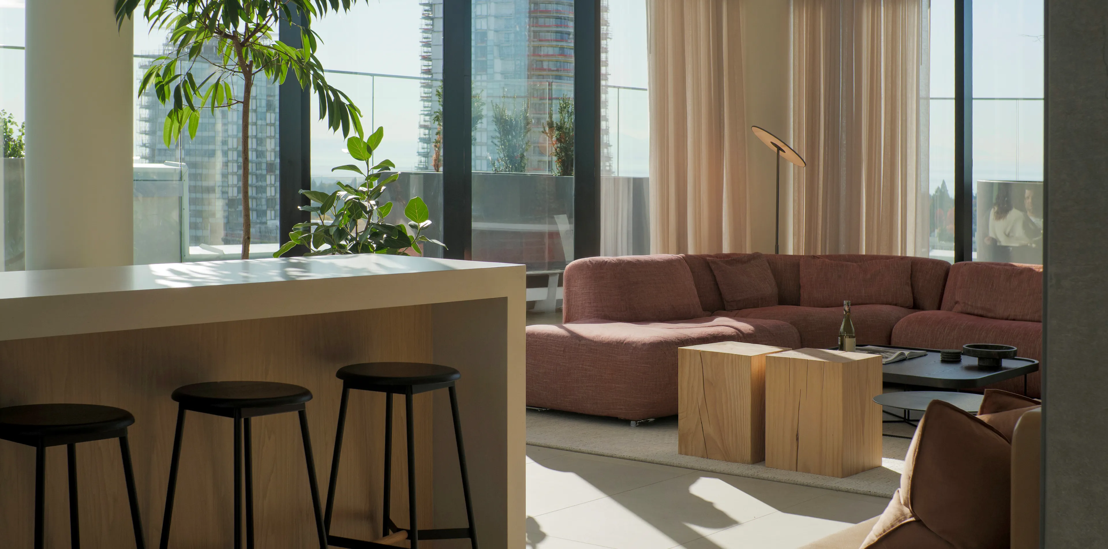 Modern living room with a large pink sectional sofa, wooden coffee tables, bar stools, and large windows offering a cityscape view.