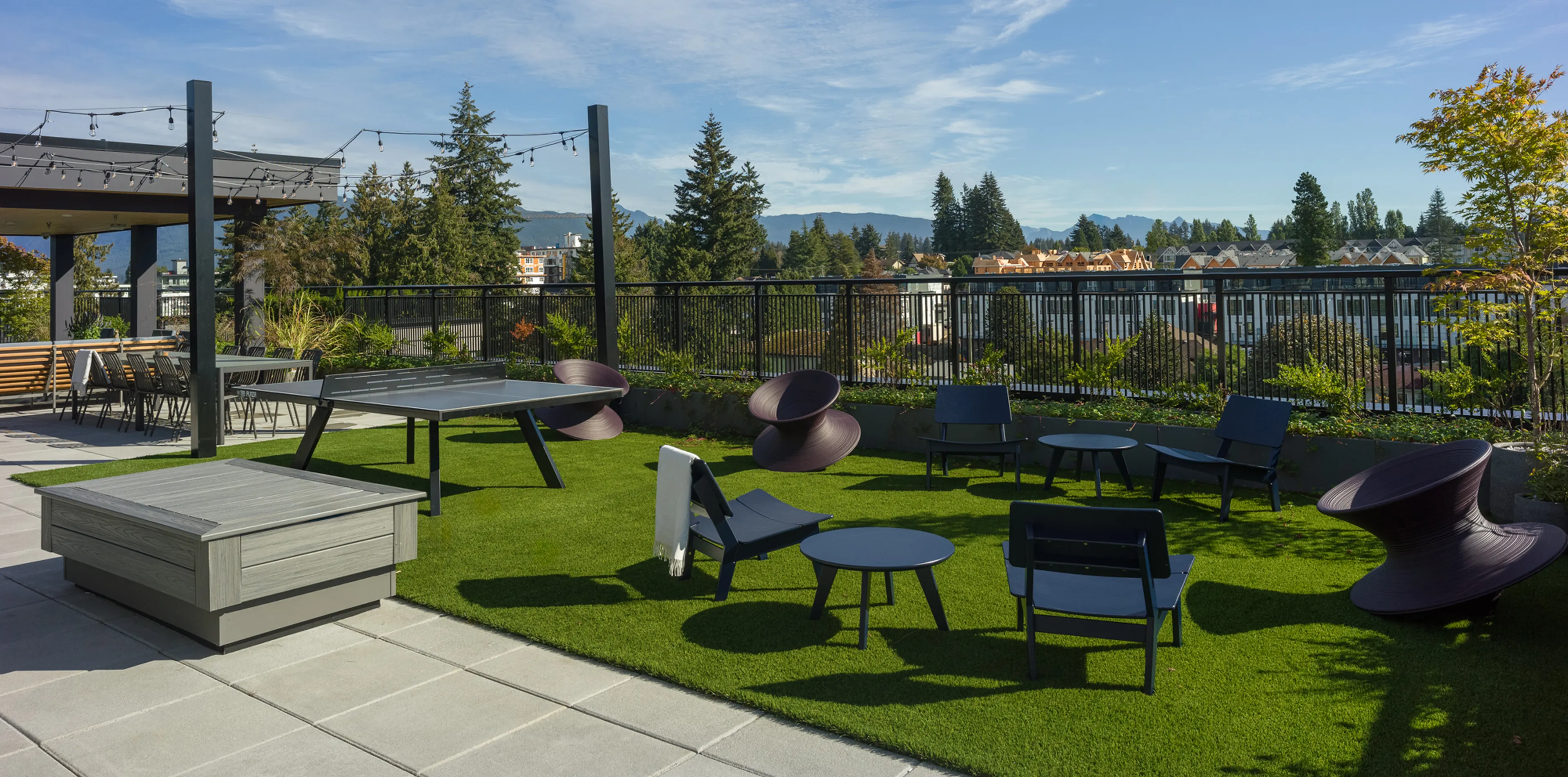 Outdoor patio with modern chairs, tables, and a ping-pong table on a grass surface, surrounded by trees and a scenic mountain view.