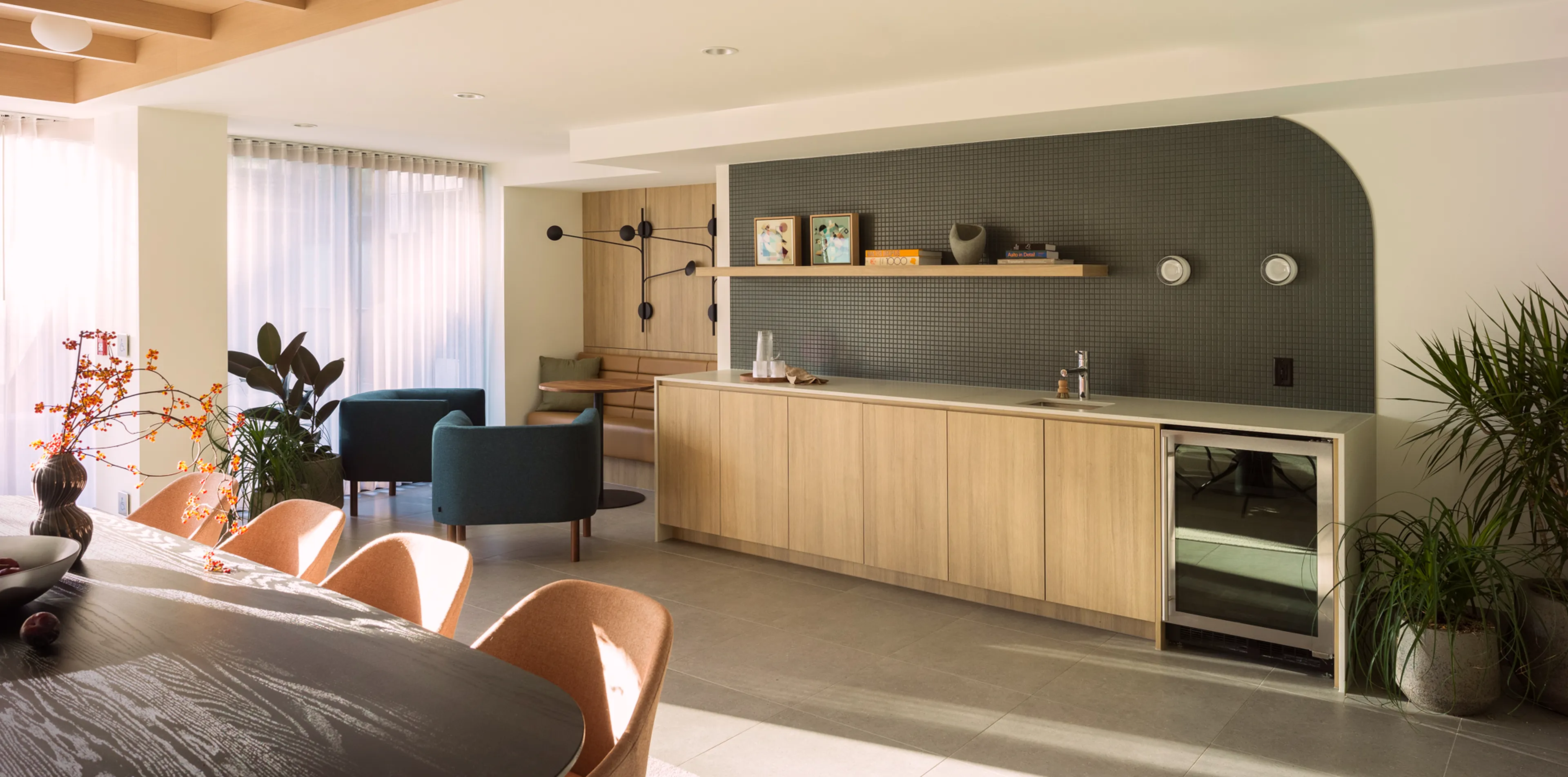 Modern living area with a wooden dining table, cushioned chairs, plants, a lounge with teal chairs, wood cabinetry, and a wall-mounted shelf.