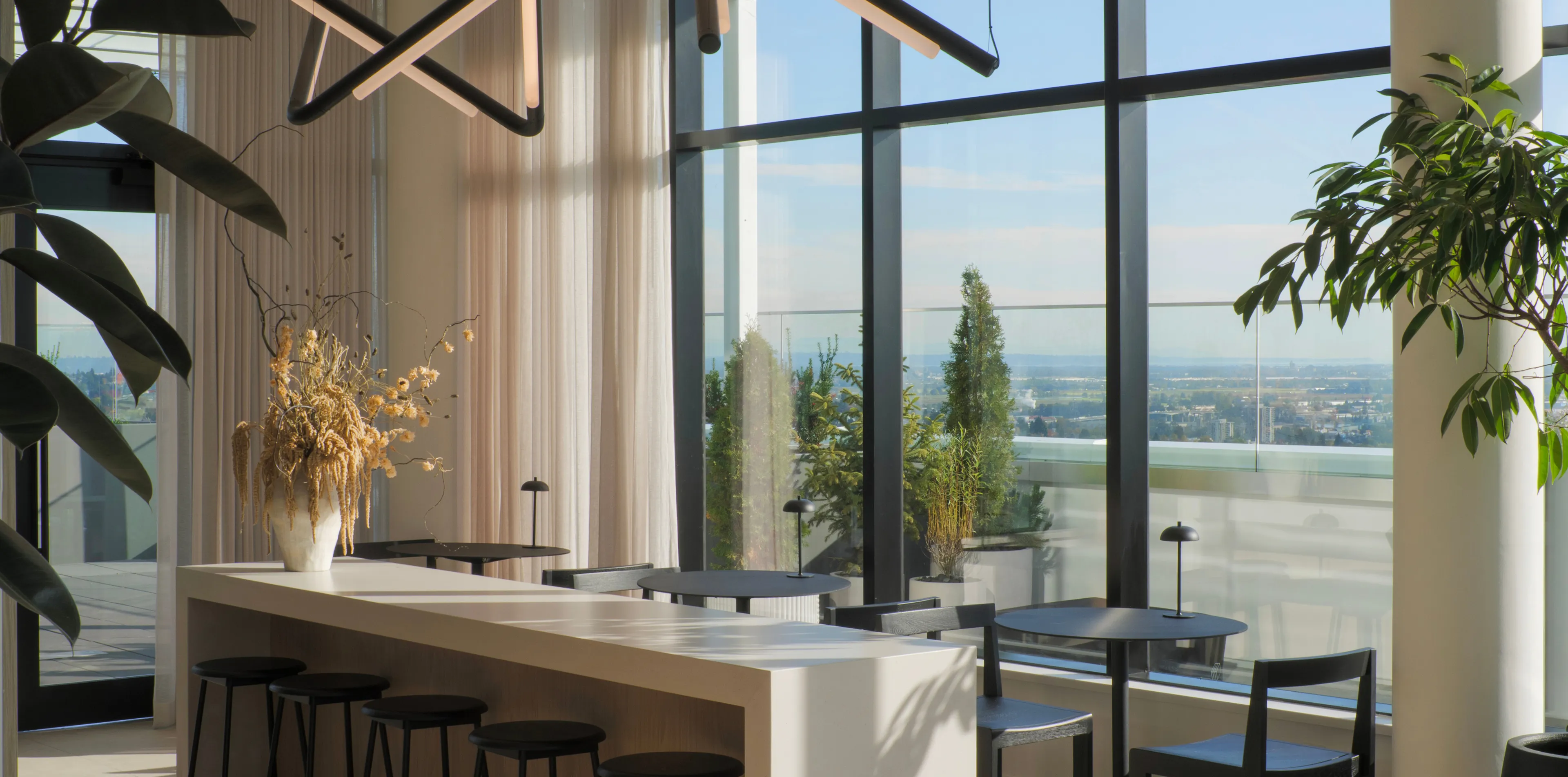 A modern indoor space with large windows, a long table with stools, black round tables and chairs, and decorative plants. Views of a city and landscape outside.