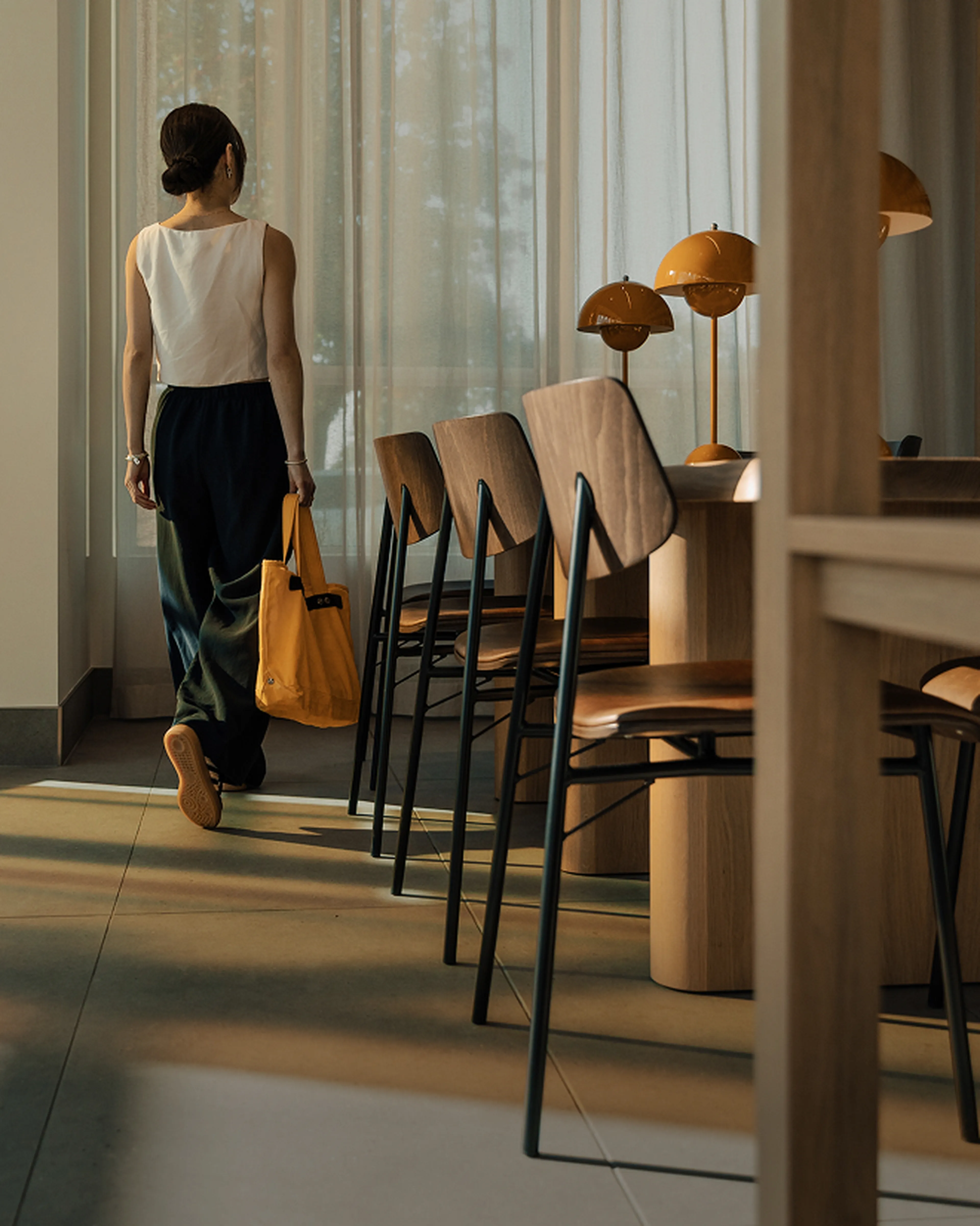 A woman in a sleeveless top and dark pants walks away, carrying a yellow tote bag in a sunlit room with wooden chairs and lamps.