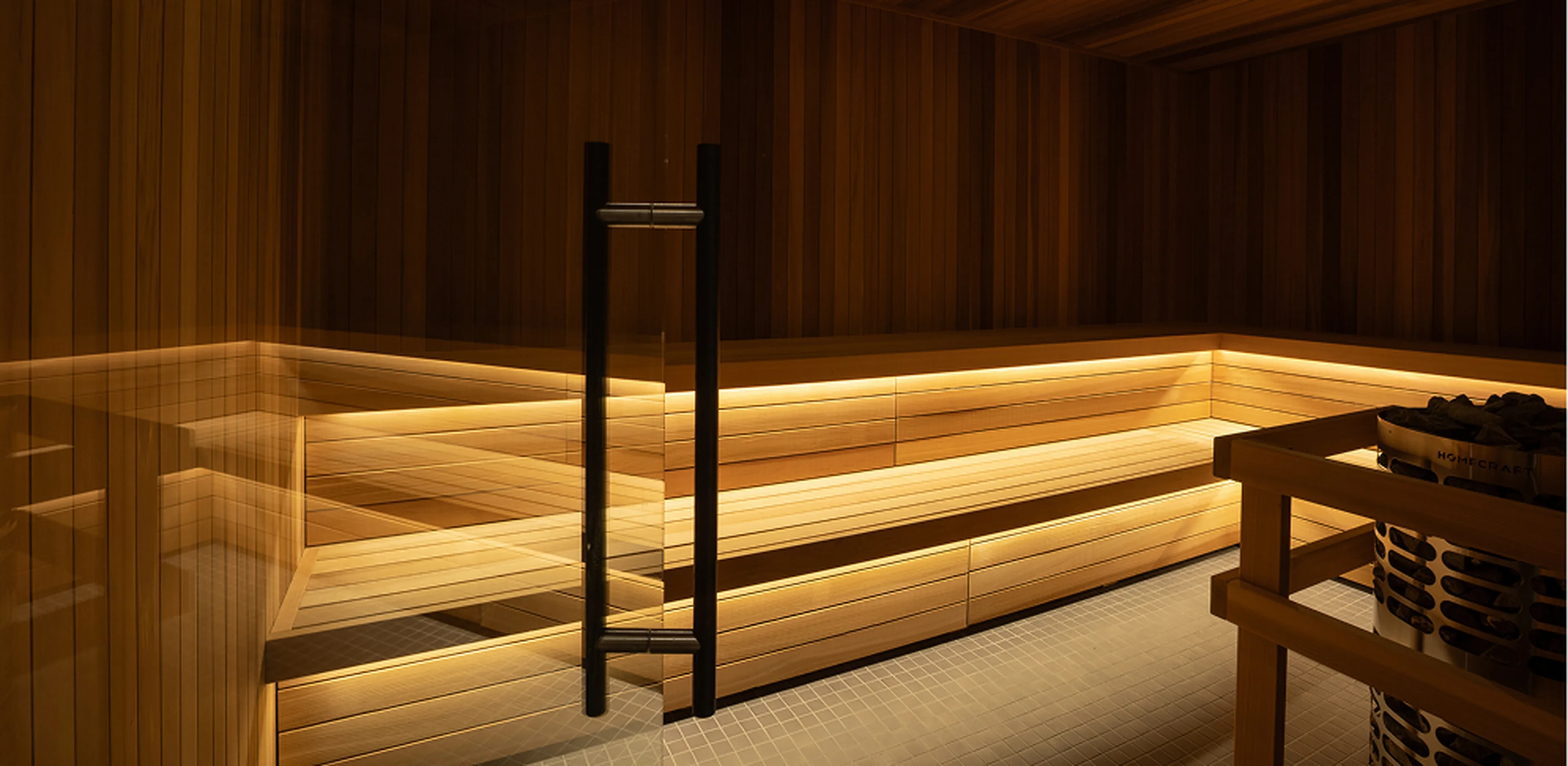 Interior of a wooden sauna with warm lighting, featuring a corner bench and a heater filled with stones.