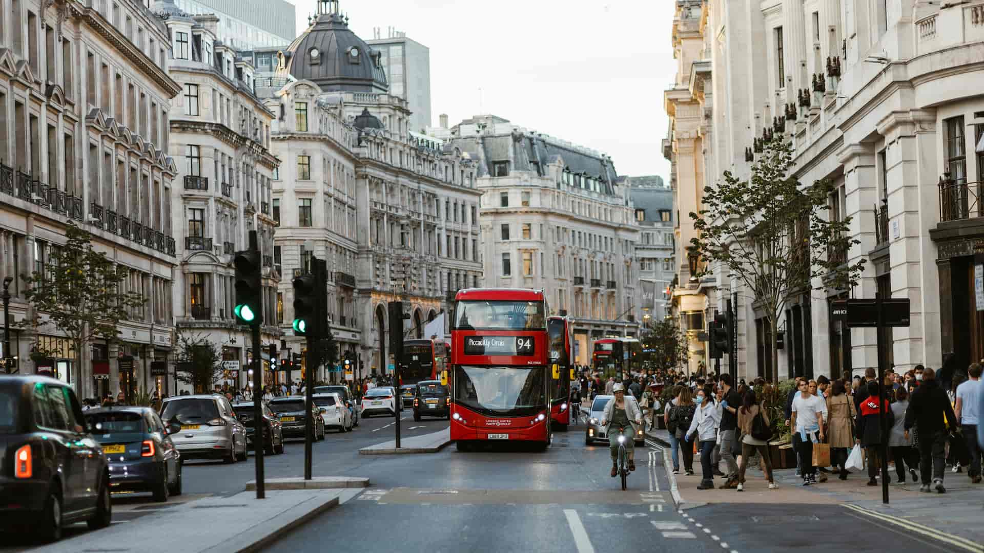 Bustling city street with red double-decker buses, cyclists, pedestrians, and classic architecture under a cloudy sky.
