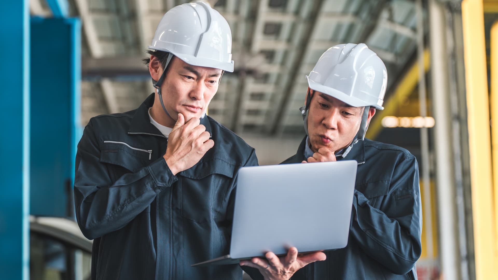 Two workers in hard hats and uniforms examine a laptop screen in an industrial setting, appearing focused and thoughtful.