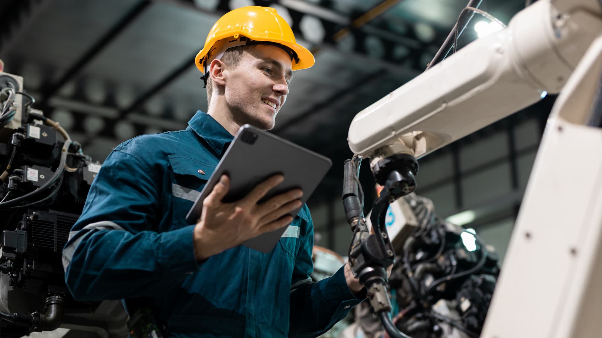 Man in a hard hat and work uniform using a tablet to operate a robotic arm in an industrial setting.