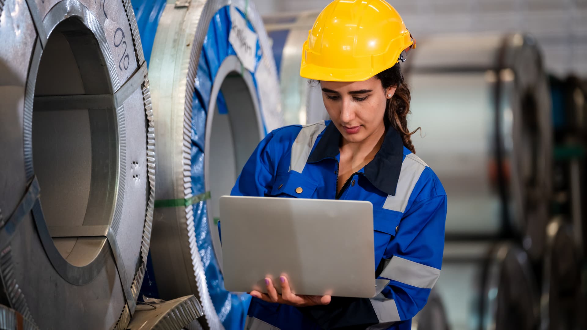 Female industrial engineer in safety helmet using laptop to inspect large metal coil rolls in manufacturing factory, ensuring quality control