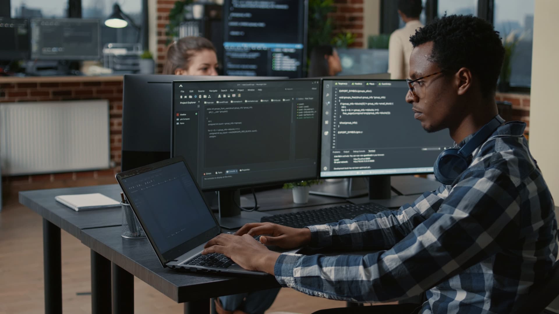 Young man in plaid shirt with headphones around his neck typing on a laptop; dual monitors display code in a modern open-plan office.