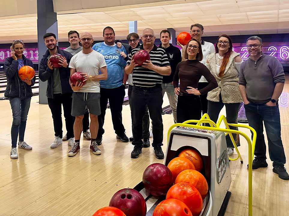 Group of people posing with bowling balls at a bowling alley, with colorful balls in a ball return machine in the foreground.