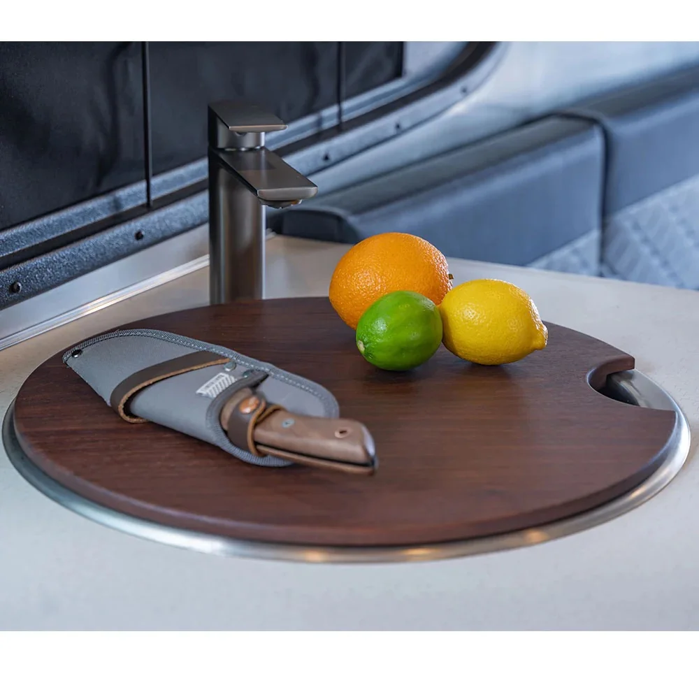 Cutting board with an orange, lemon, lime, and a sheathed knife near a metal faucet in a modern kitchen setting.