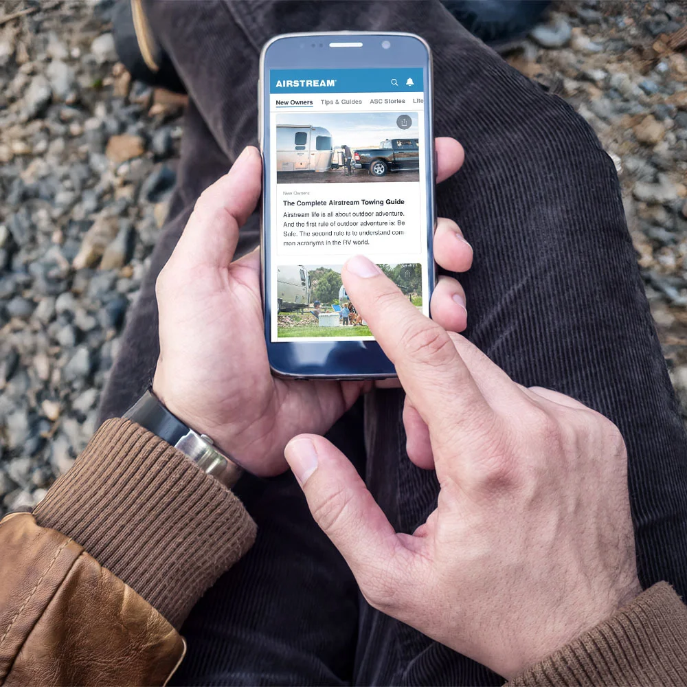 Person wearing a brown jacket and dark pants browsing a guide on a smartphone while sitting on gravel.
