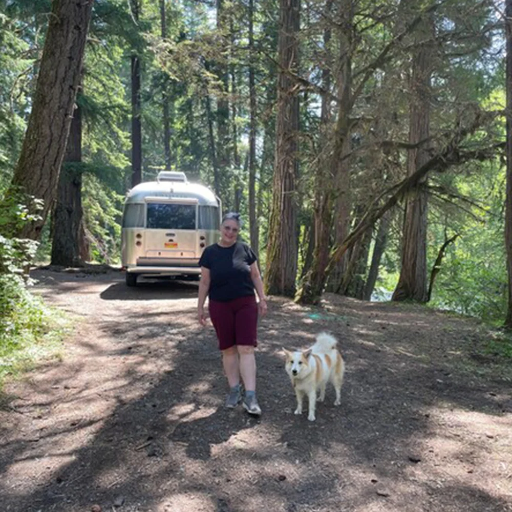 A person walks with a dog on a wooded path near a silver travel trailer parked among tall trees.