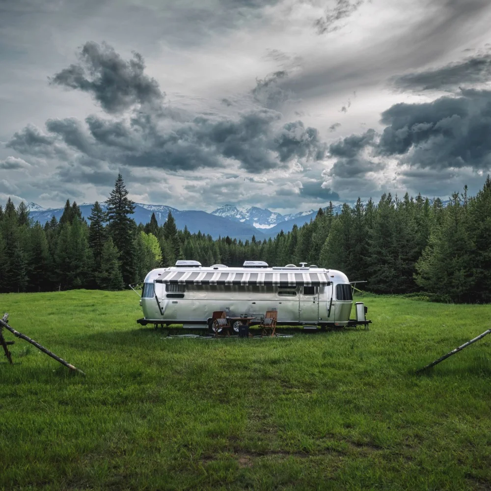 A silver trailer sits on a lush green field, surrounded by pine trees and mountains under a dramatic cloudy sky.