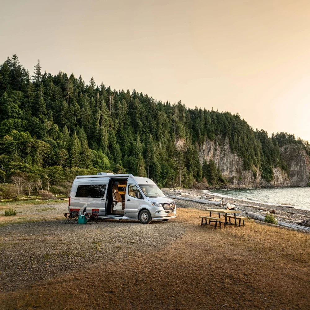 Camper van parked by a forested cliff and beach, with camping chairs and a picnic table nearby under a serene, pale orange sky.