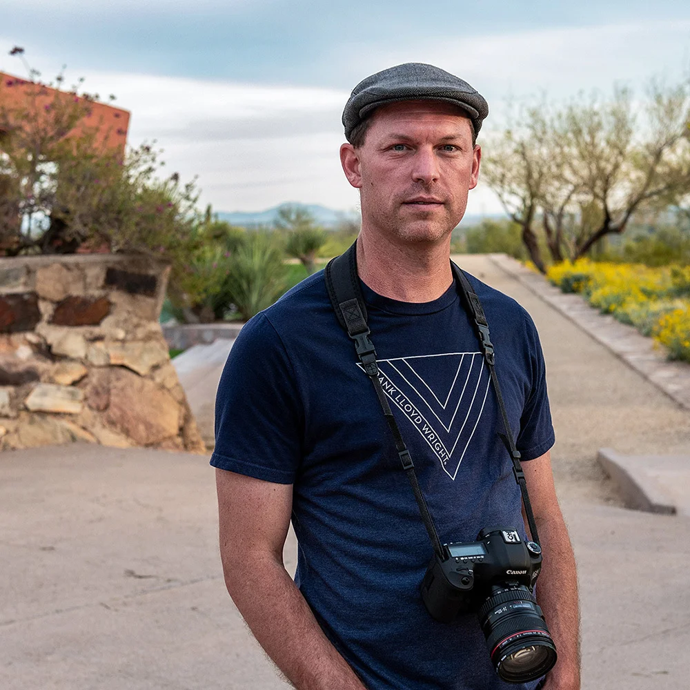 Person with a flat cap and camera around their neck stands on a path surrounded by trees and flowers under a cloudy sky.
