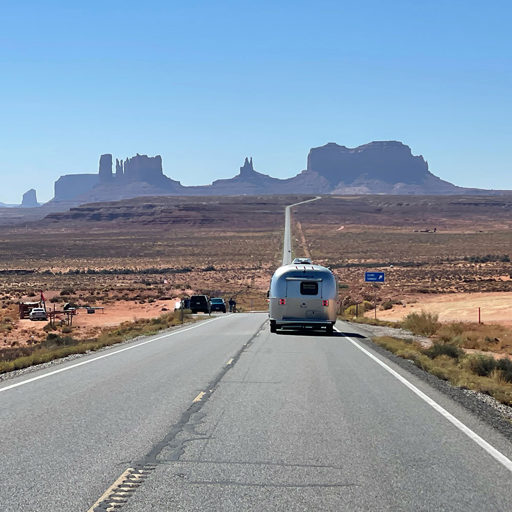 Airstream trailer traveling down a long, straight road towards distant mesas and buttes in a desert landscape under a clear blue sky.